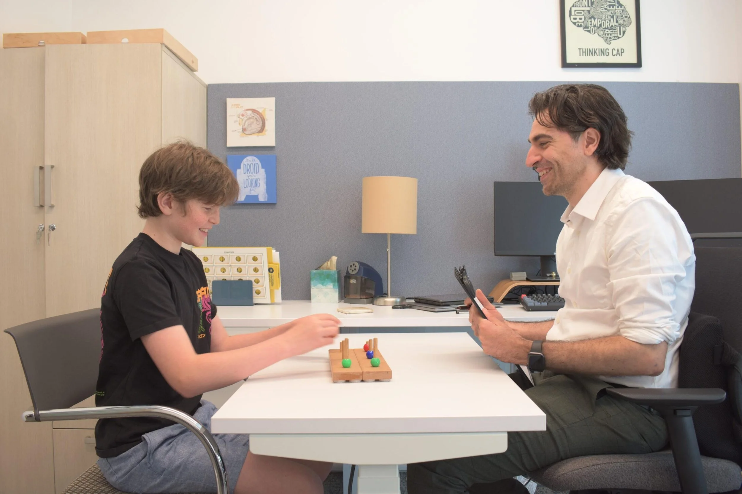 A young boy and an adult man smiling and playing a board game together at a desk in an office or classroom setting. The boy is seated, and the man is standing, holding a phone. There are posters and office supplies on the wall and desk.