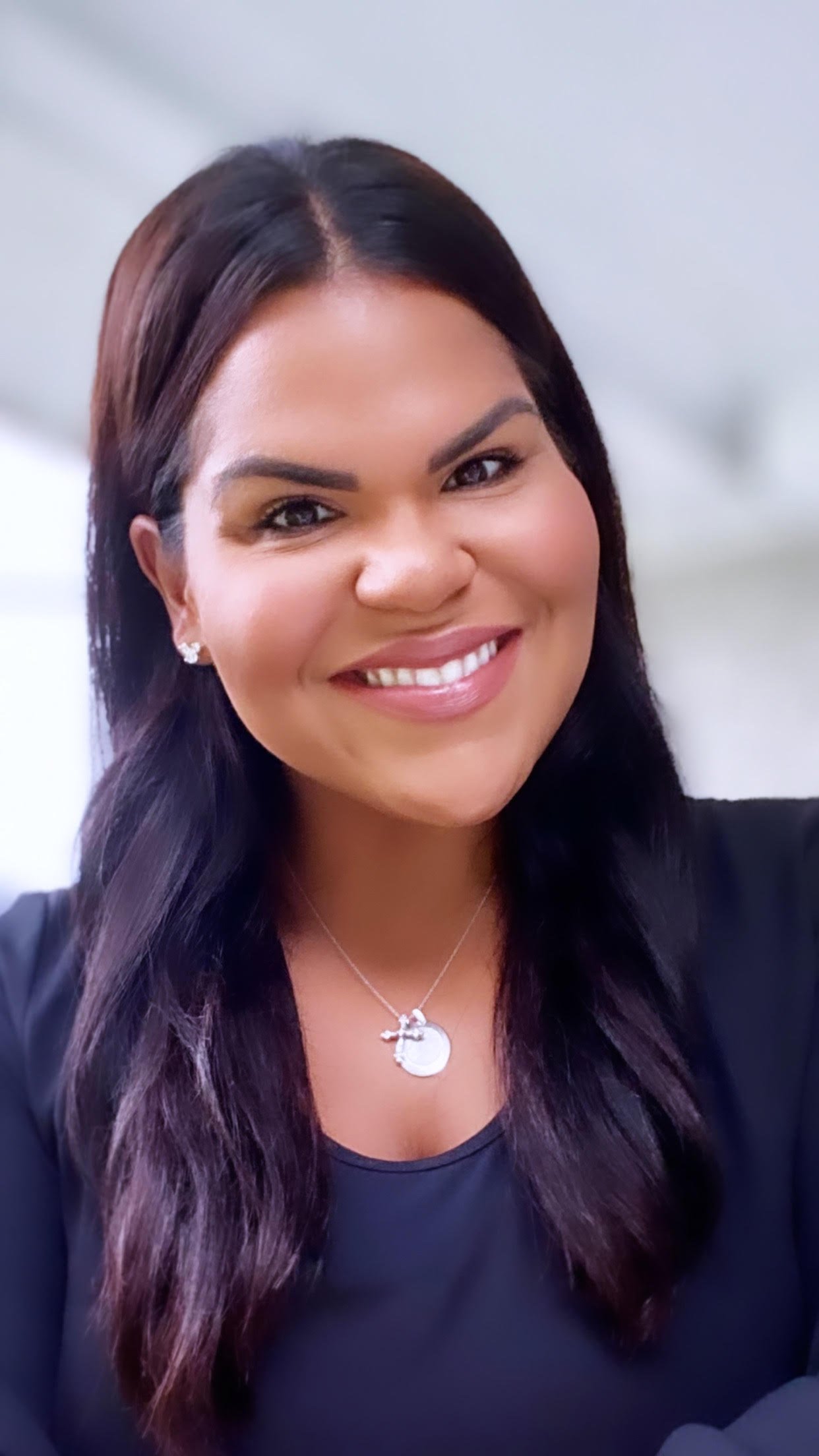 A woman with long dark hair, smiling, wearing a black top, earrings, and a necklace with a moon pendant.