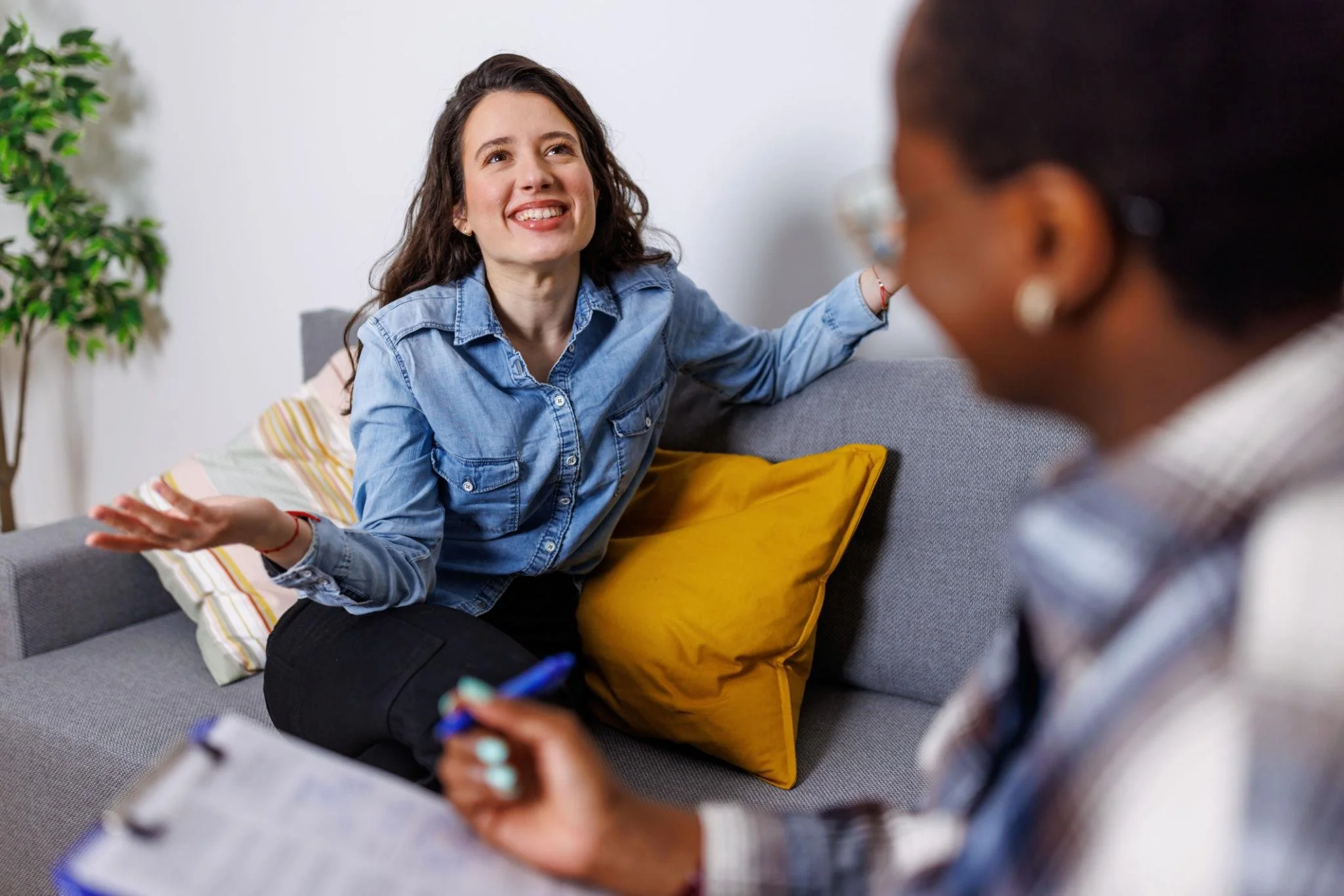 A woman with long brown hair and a denim shirt smiling and talking to another woman who is taking notes.