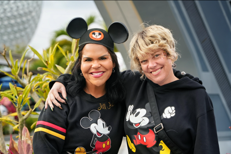 Two women smiling, one wearing a Mickey Mouse beanie and both wearing Mickey Mouse shirts, standing outdoors with greenery.
