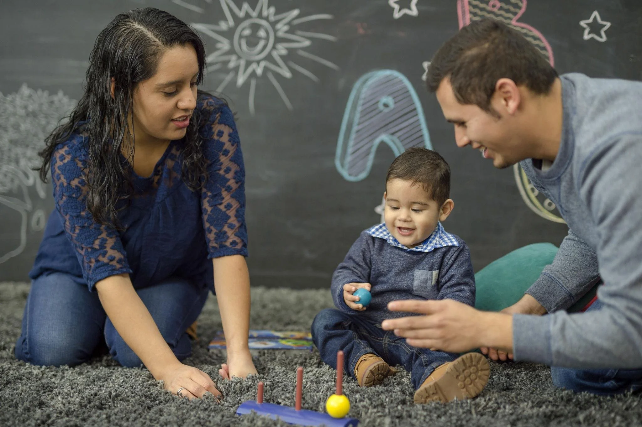 A group of three people, two adults and one child, sit on a carpeted floor playing with small, colorful game pieces in front of a black chalkboard with drawings of a smiling sun, stars, clouds, and a rainbow.