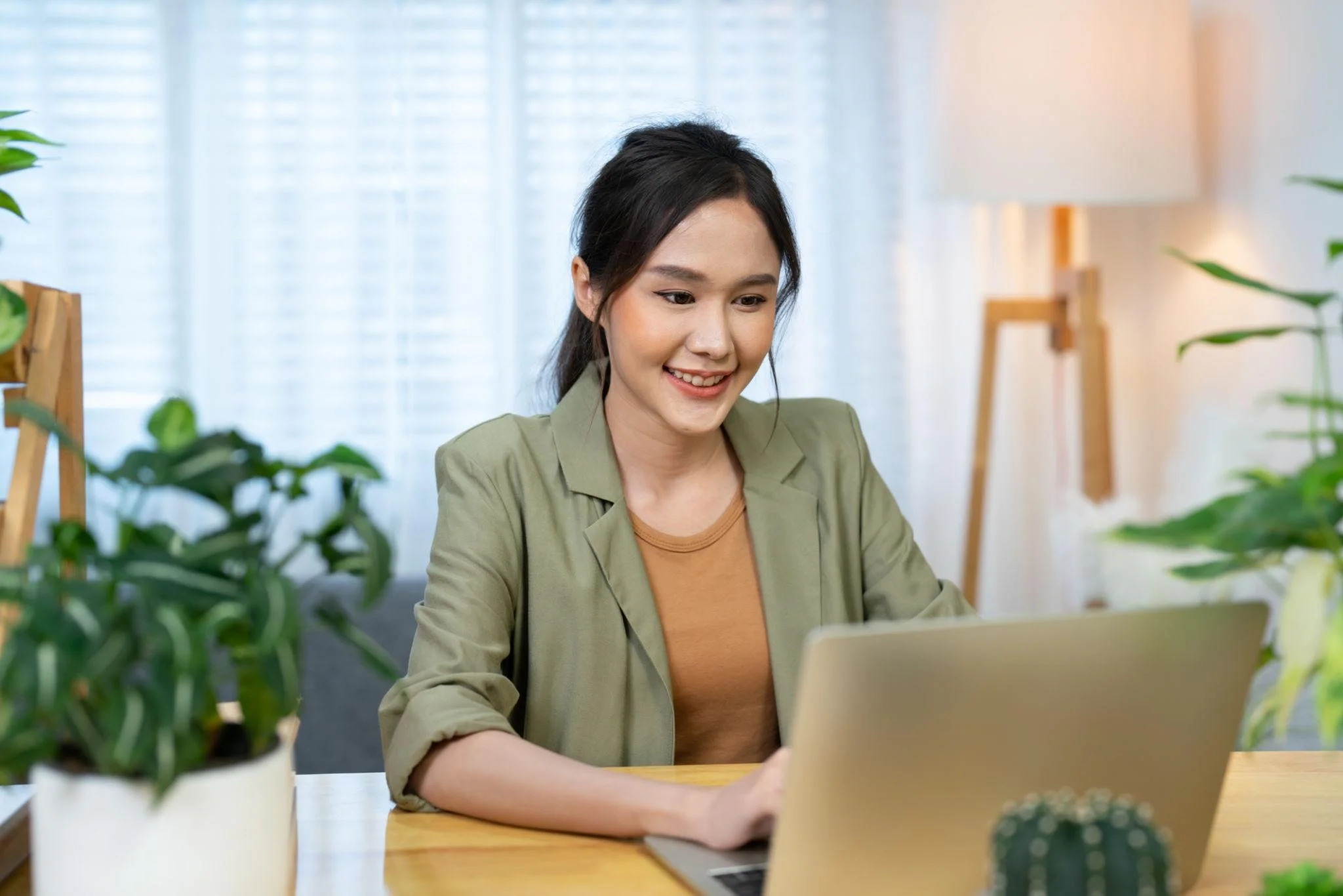 A young woman with dark hair smiling and working on a laptop at a desk surrounded by potted plants in a bright room with white curtains.