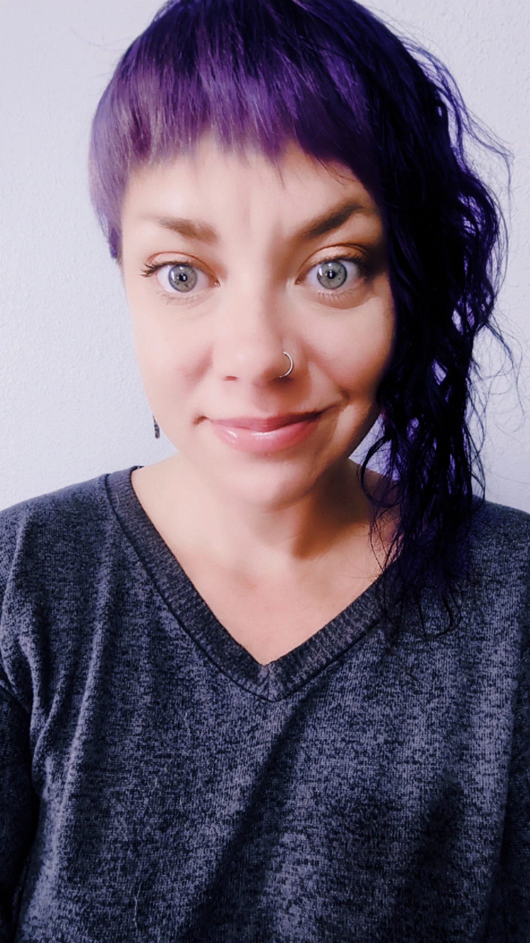 A woman with short purple and black hair, blue eyes, pierced nose, smiling, wearing a dark gray v-neck shirt, against a plain light-colored wall.
