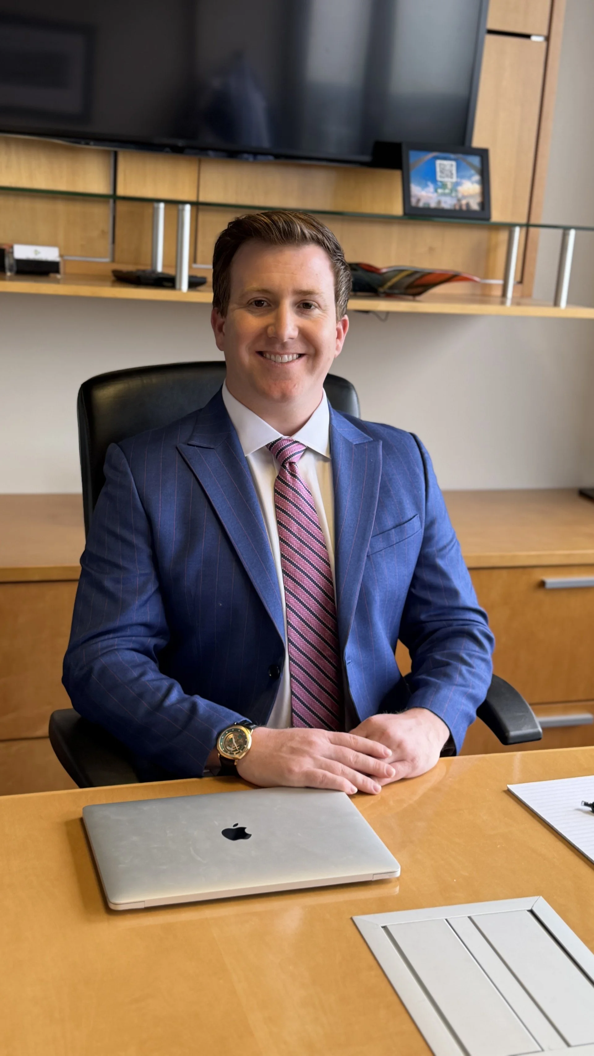 A man in a blue suit and pink striped tie sitting at a desk in an office, smiling at the camera, with a laptop and notepad on the desk.