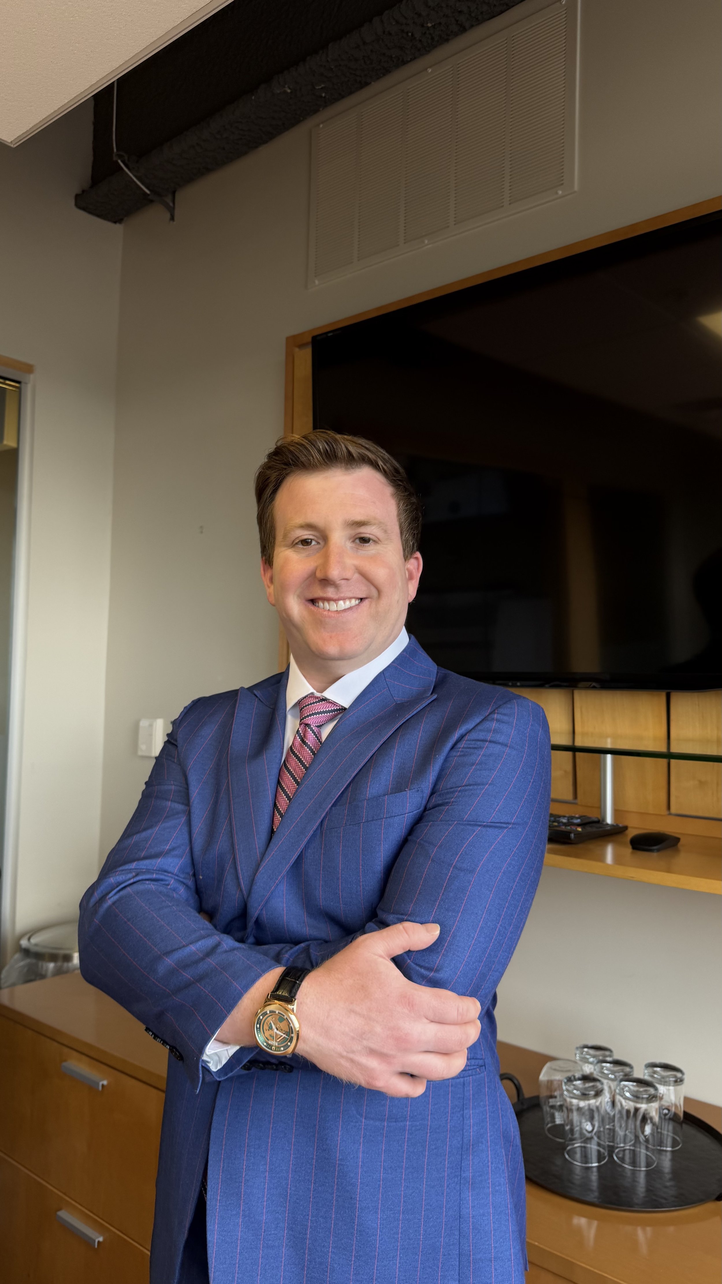 A smiling man wearing a blue pinstripe suit, pink tie, white shirt, and a watch, standing with arms crossed in an office or conference room.