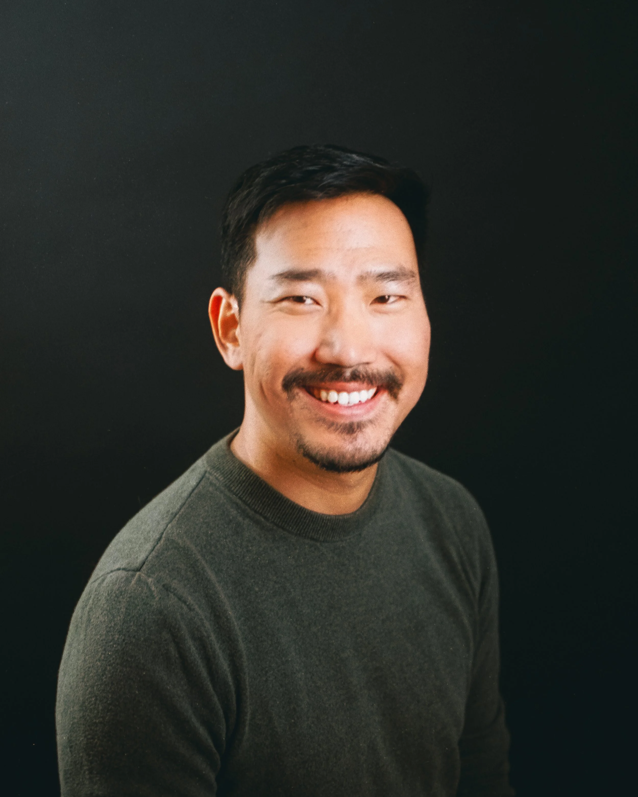 A smiling man with black hair, a short beard, and a mustache, wearing a dark green sweater against a black background.