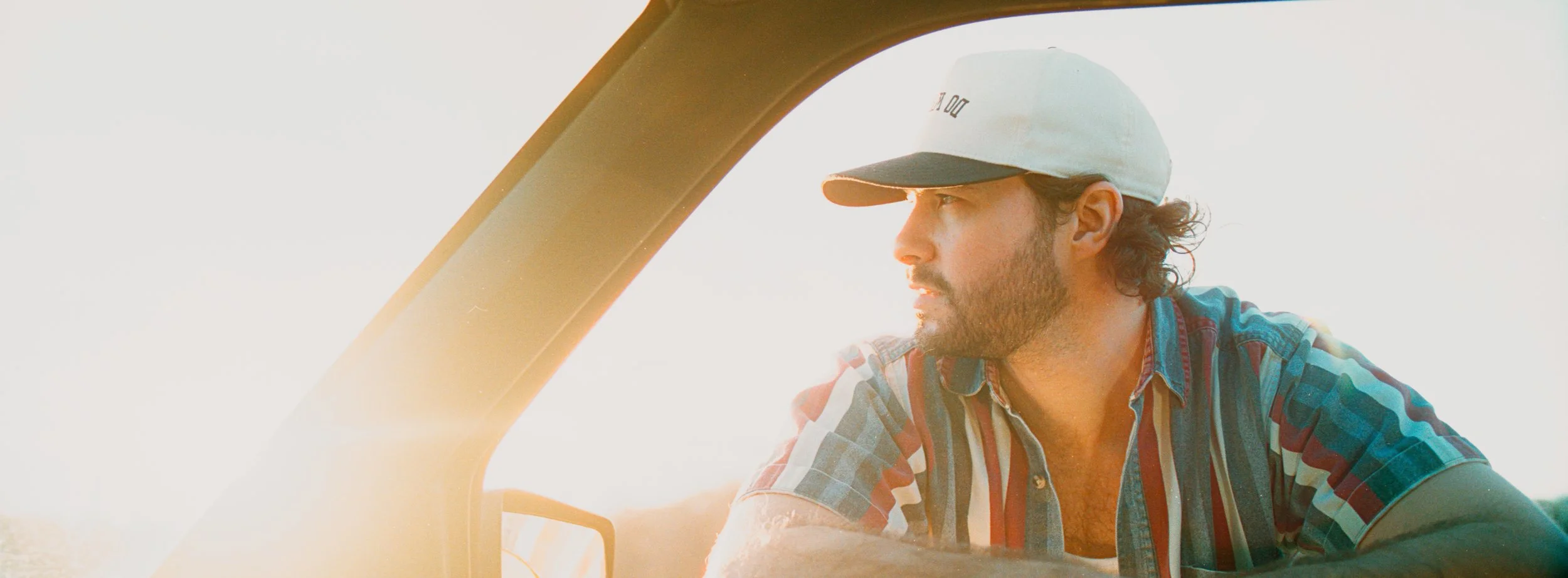 A man with a beard and curly hair, wearing a beige baseball cap and a plaid shirt, looking to the right while leaning out of a vehicle window at sunset.