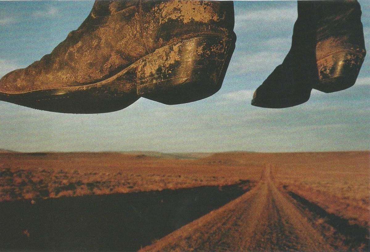 The image shows a surreal scene where large, weathered boots hover above a dirt road in a barren landscape.