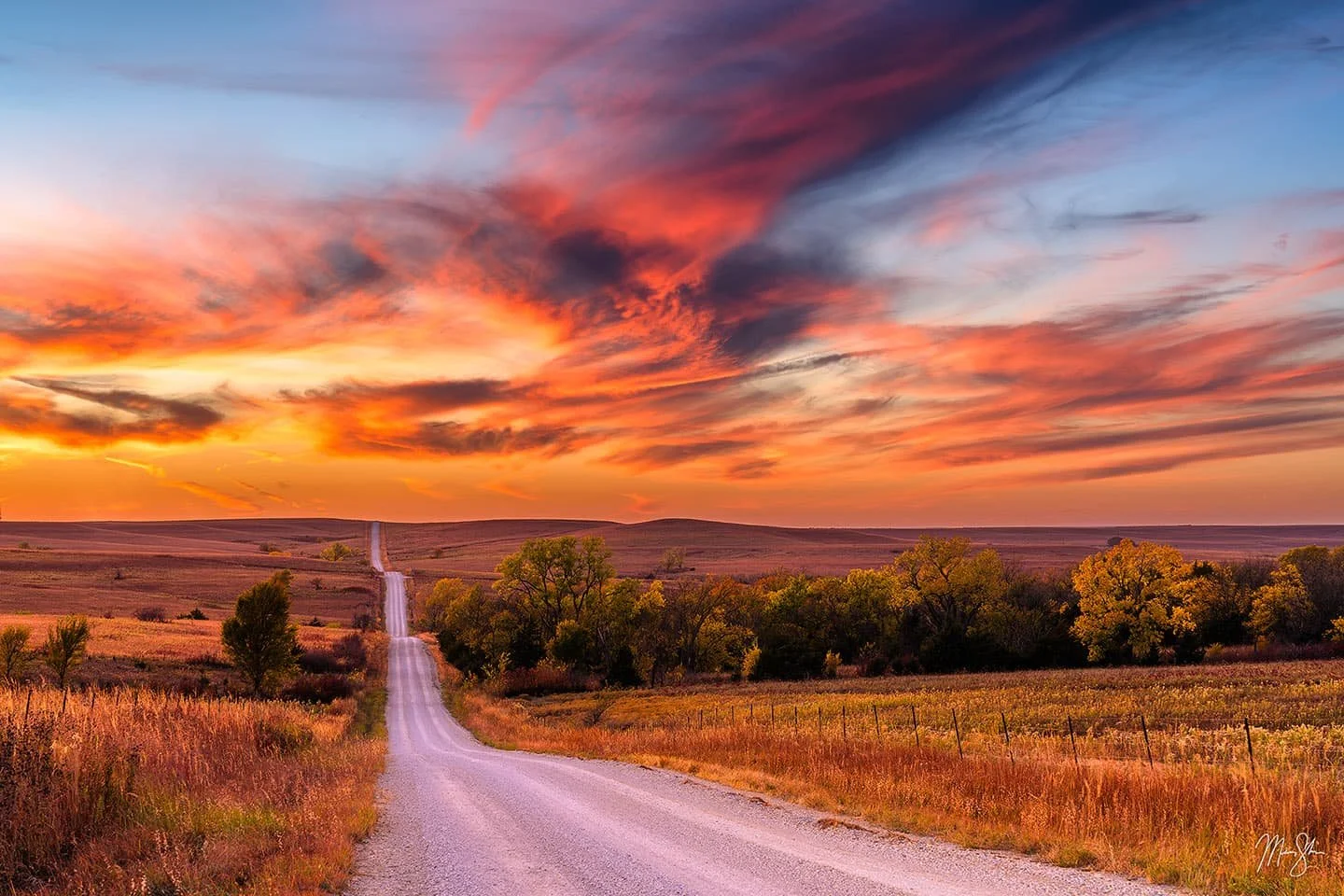 A scenic landscape with a dirt road leading into the horizon, surrounded by trees and fields during sunset, with colorful clouds in the sky.
