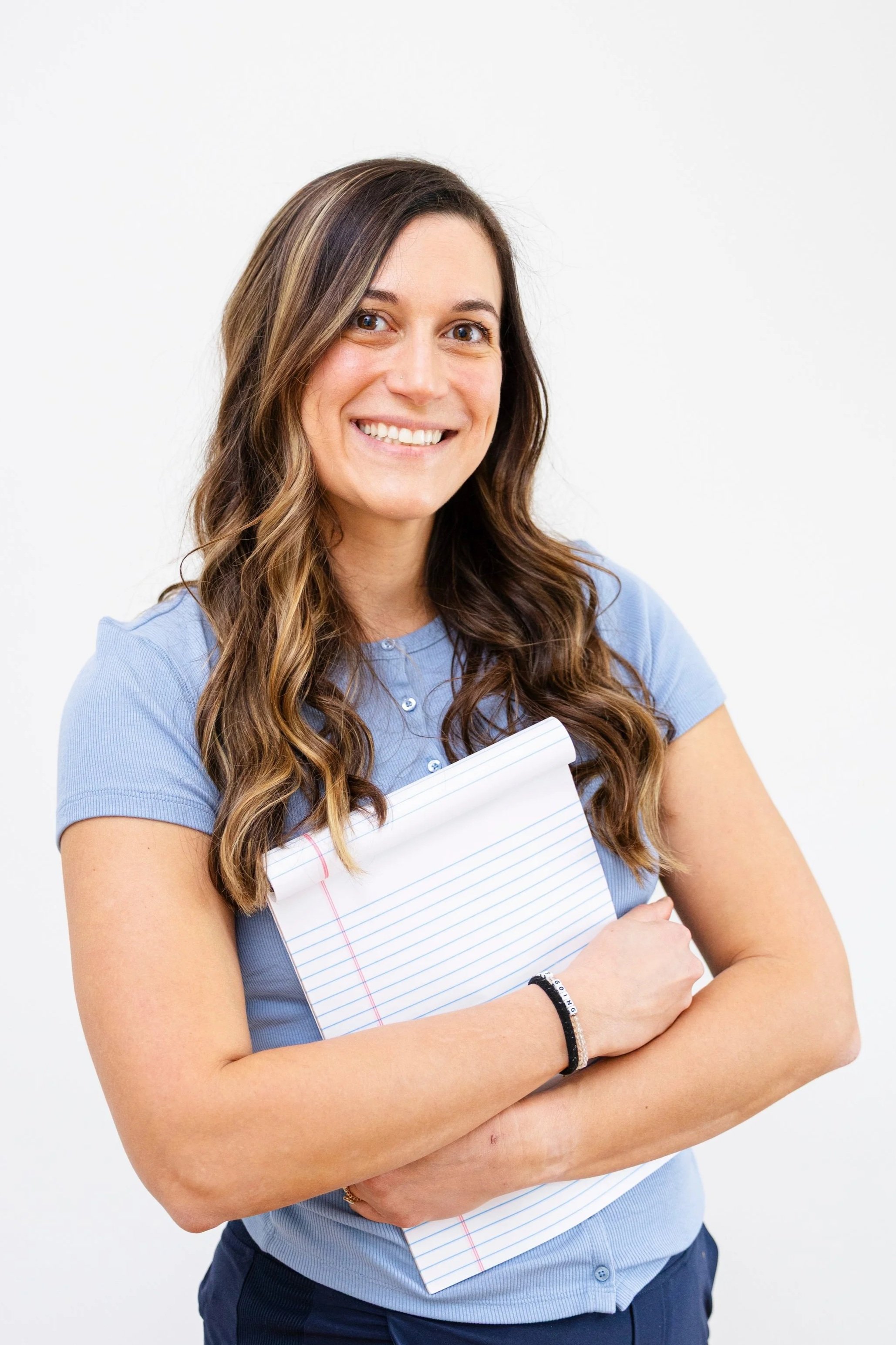 A woman with long brown wavy hair, smiling, wearing a light blue polo shirt, holding a spiral notebook, standing against a plain white background.
