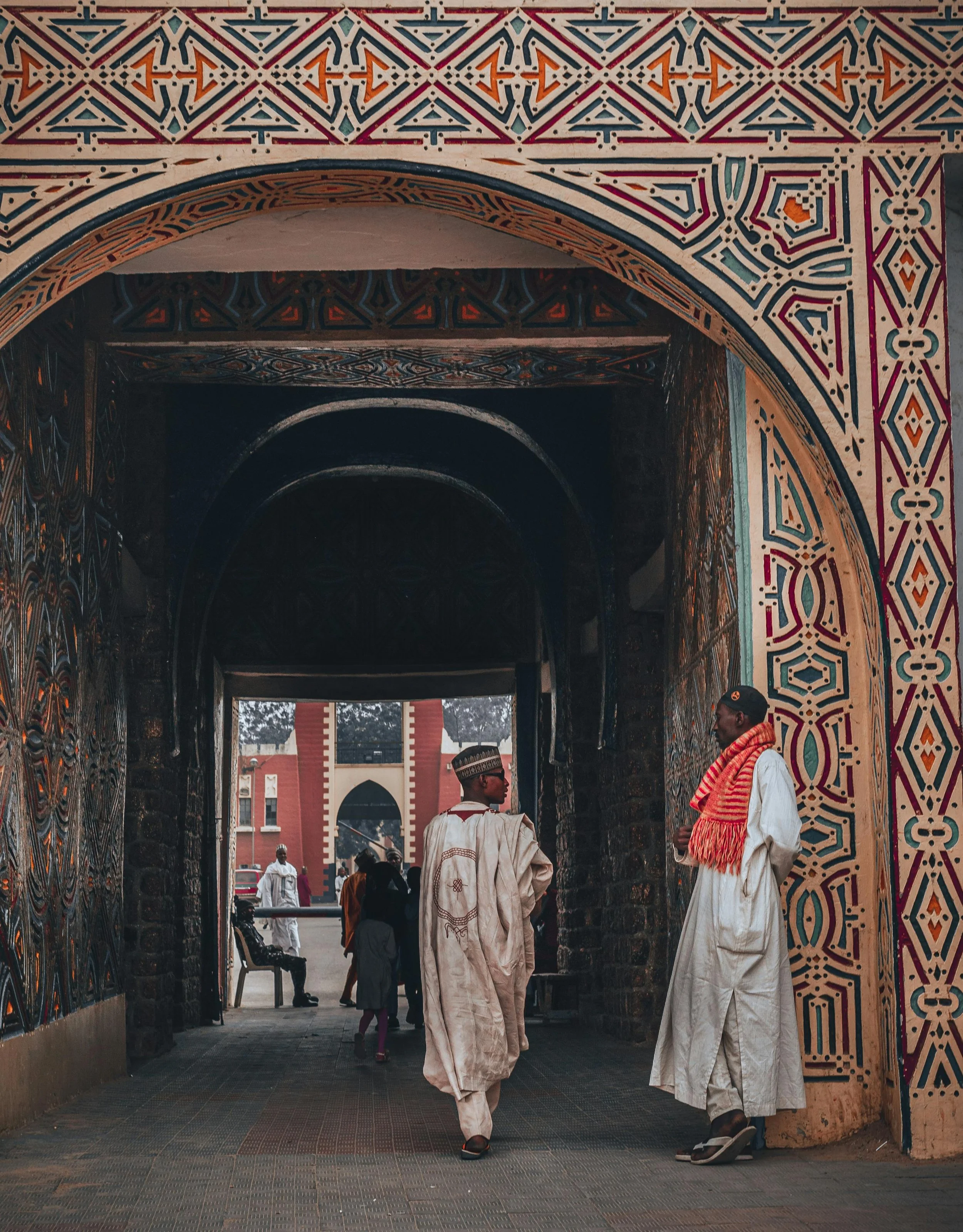 Two men wearing traditional African attire standing in a decorated archway, with people and a red brick building visible in the background.
