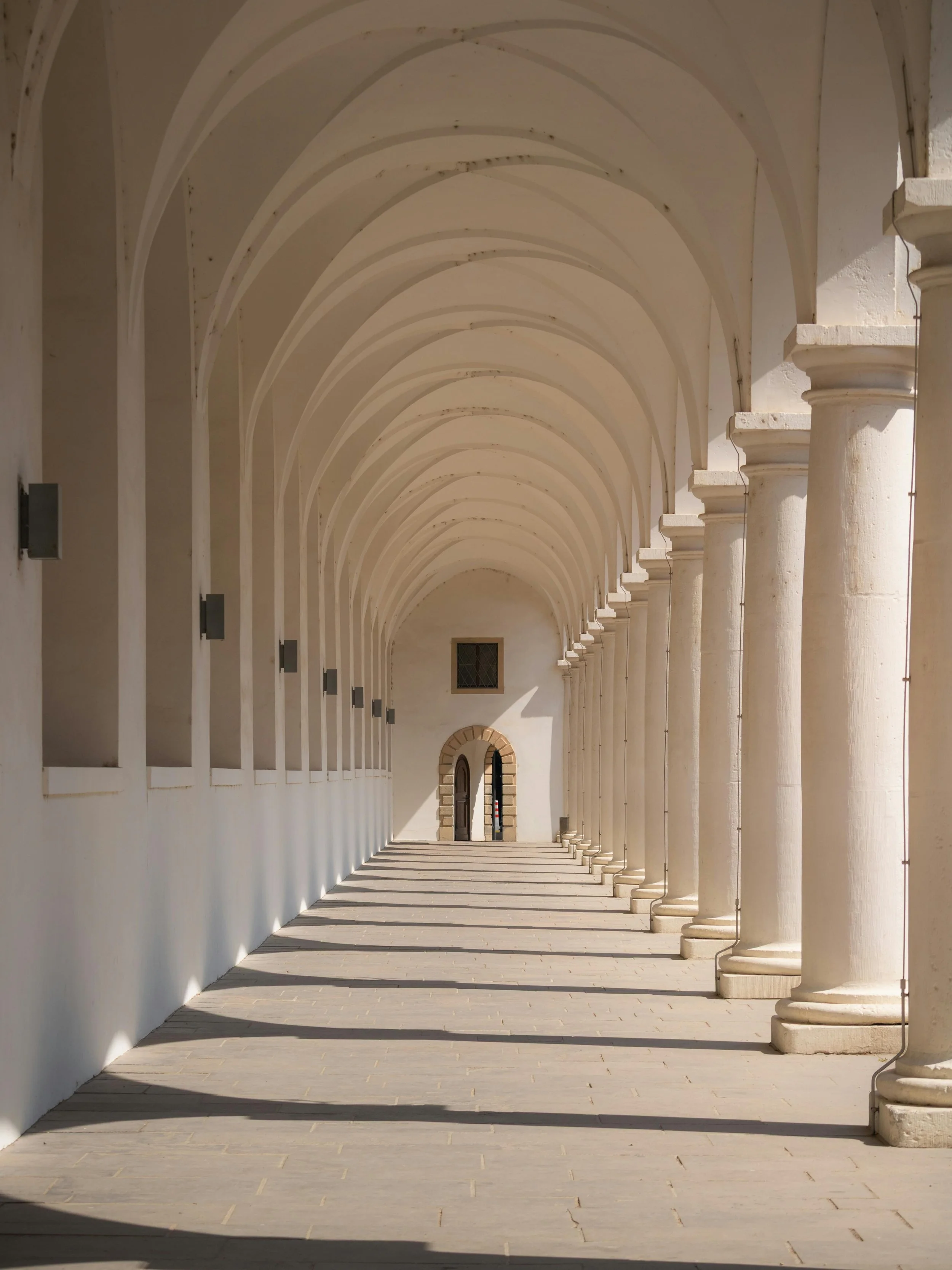 A corridor with white arches and columns, casting shadows on the floor, leading to a door at the end.