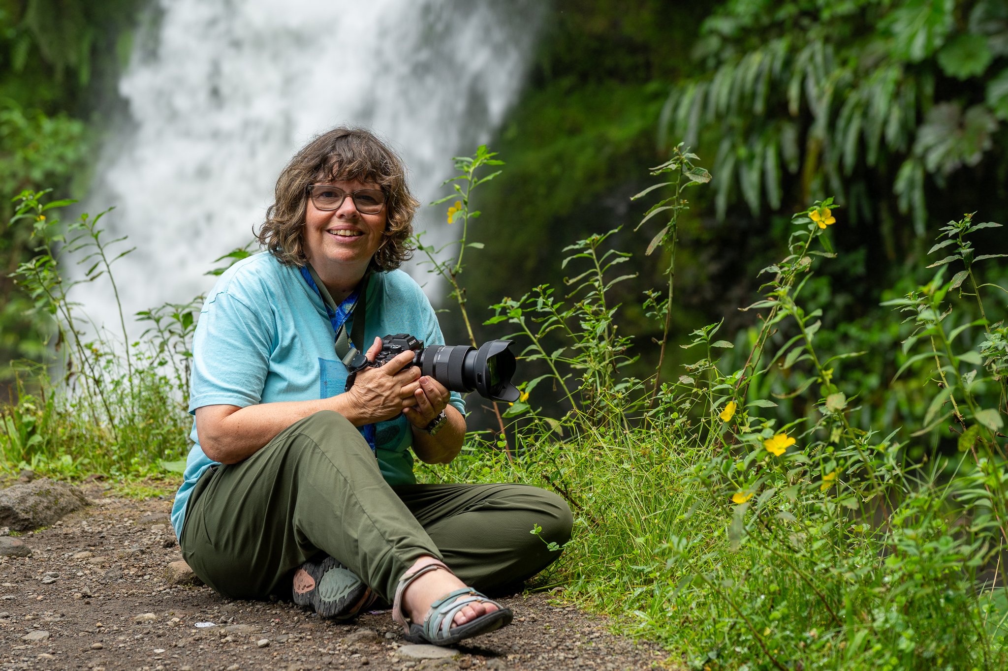 Linda Detwiler Burner sitting on the ground holding a camera, smiling, near a waterfall in a lush green forest.