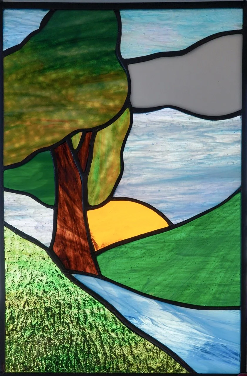 Stained glass window depicting a landscape with a green tree, a yellow setting sun, a blue river, and a grassy field.