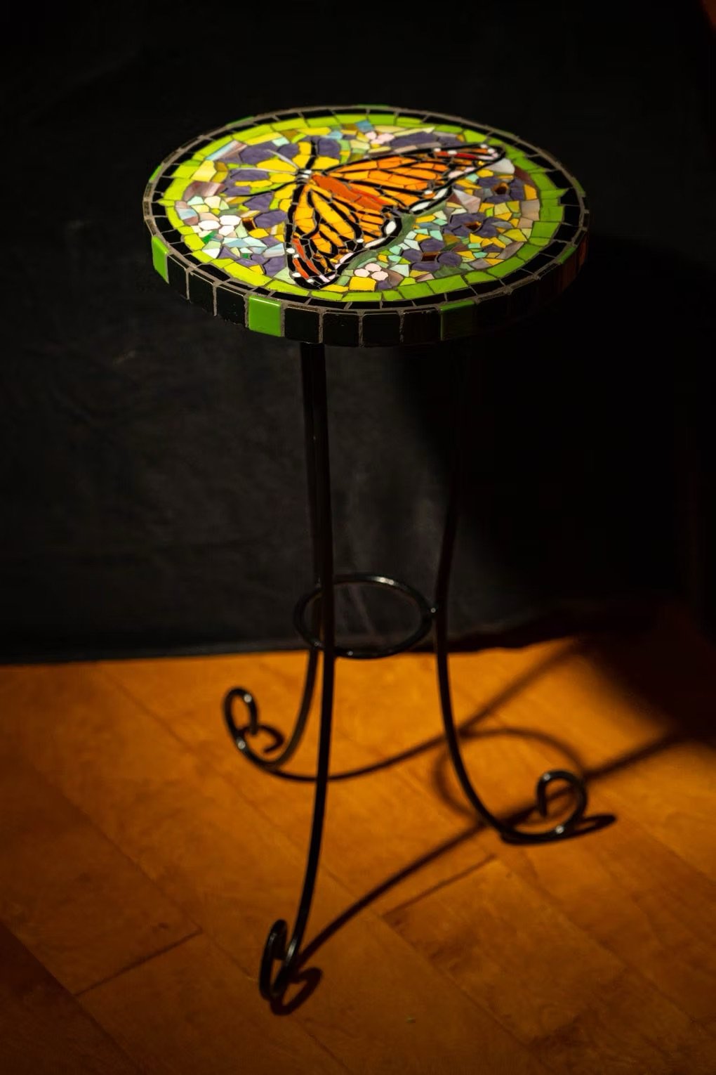 Handcrafted mosaic butterfly table by Linda Detwiler Burner, supported by a black metal stand with decorative curled legs, placed on a wooden floor.