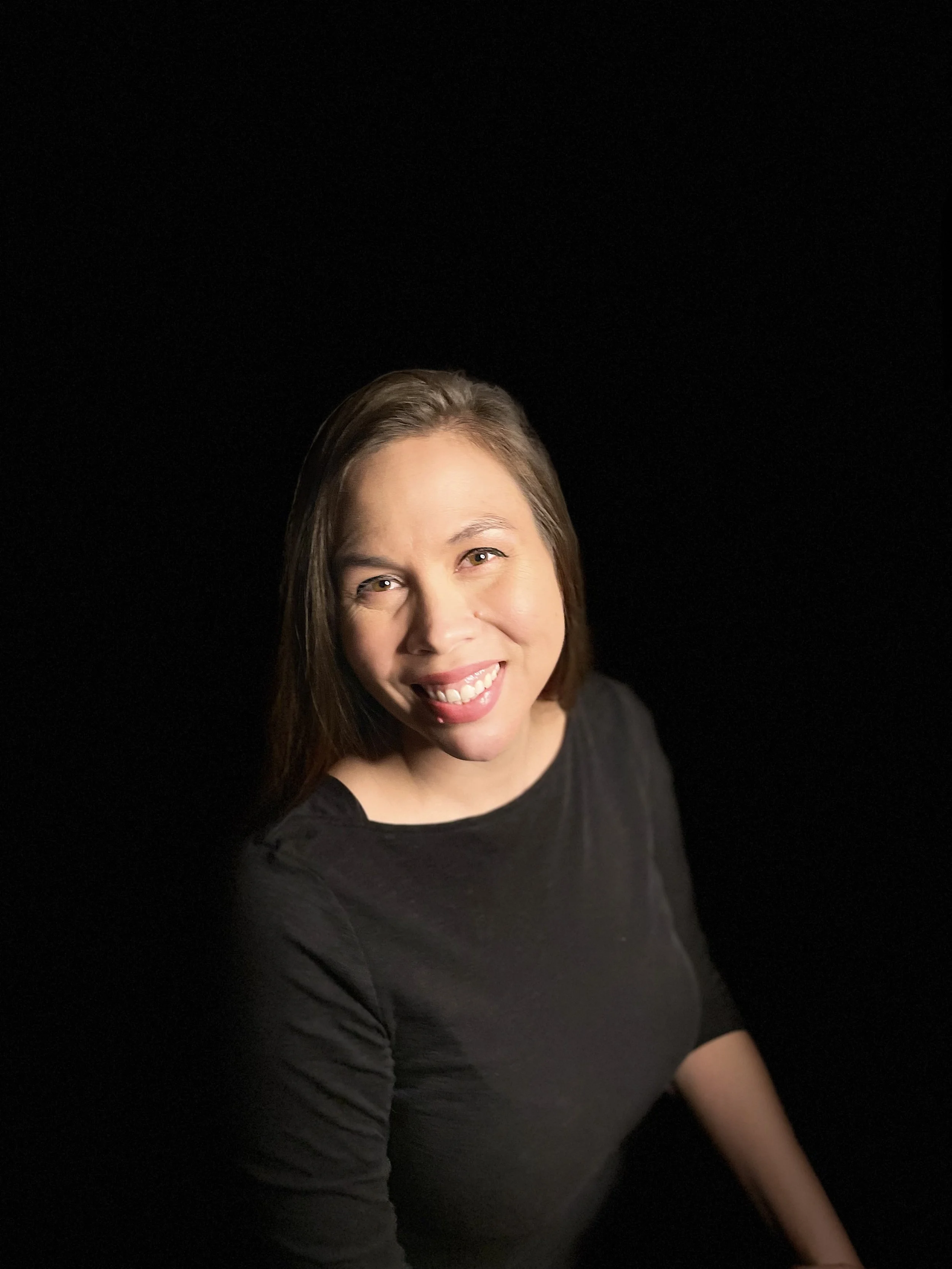 A woman with brown hair smiling at the camera, wearing a black top, against a plain black background.