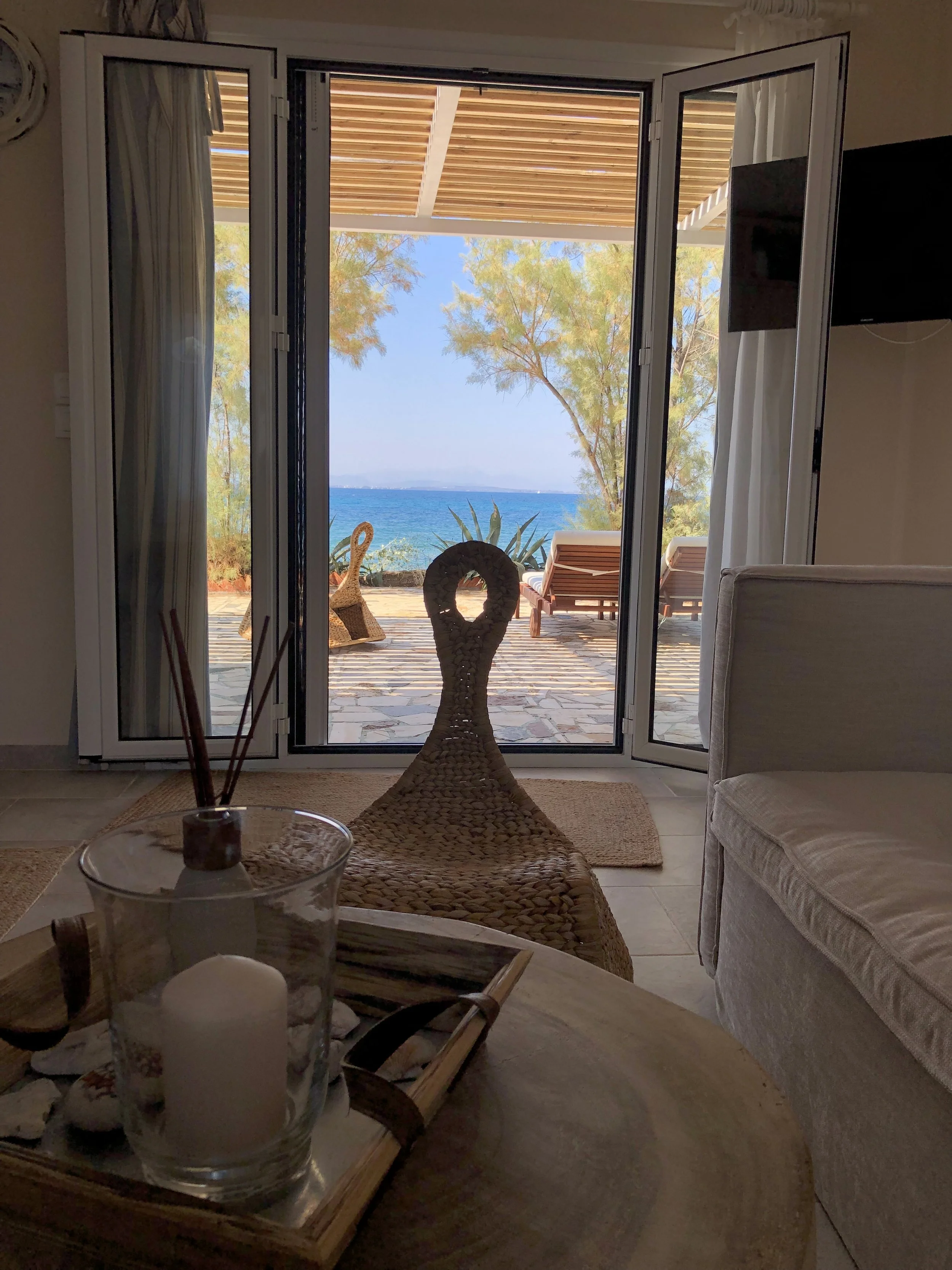 Living room with open glass doors leading to an outdoor patio overlooking the ocean, with lounge chairs, trees, and a clear blue sky in the background.