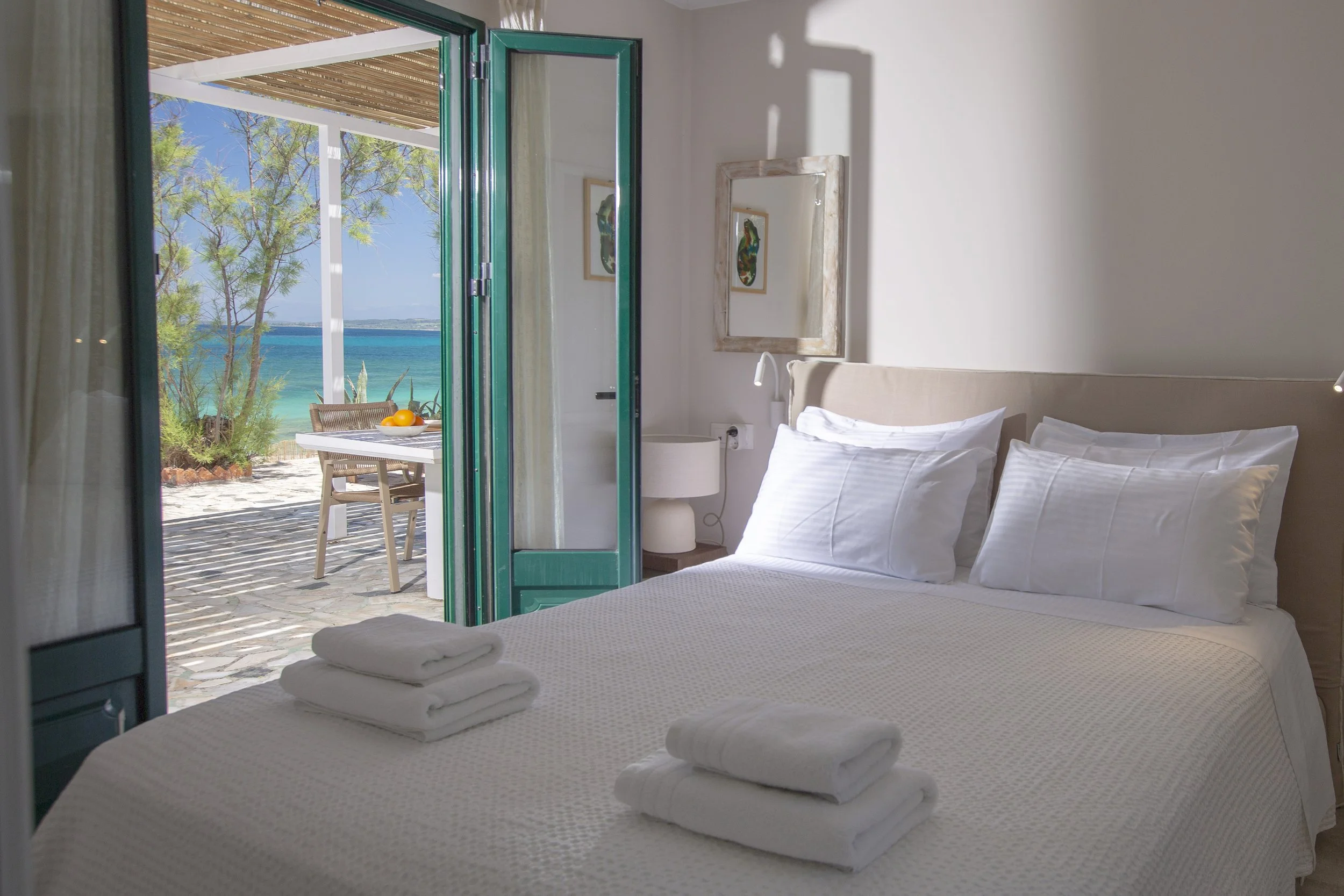 A bedroom with a large bed made up with white linens and towels folded on top, opening to a balcony with a view of the beach, trees, and blue ocean, with framed artwork on the wall.