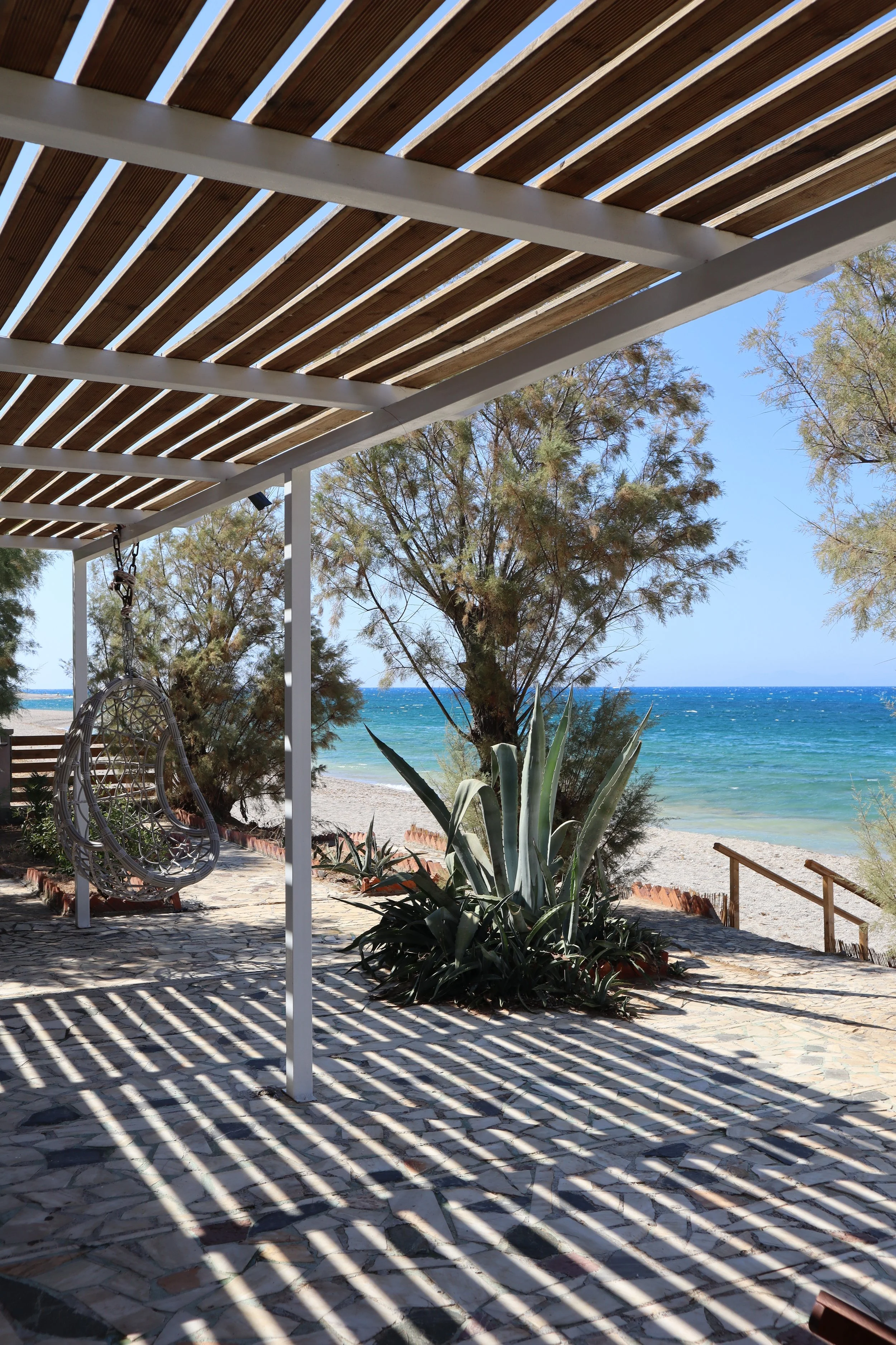Beachside patio with a shaded pergola, a hanging egg chair, sandy beach, ocean waves, and trees.