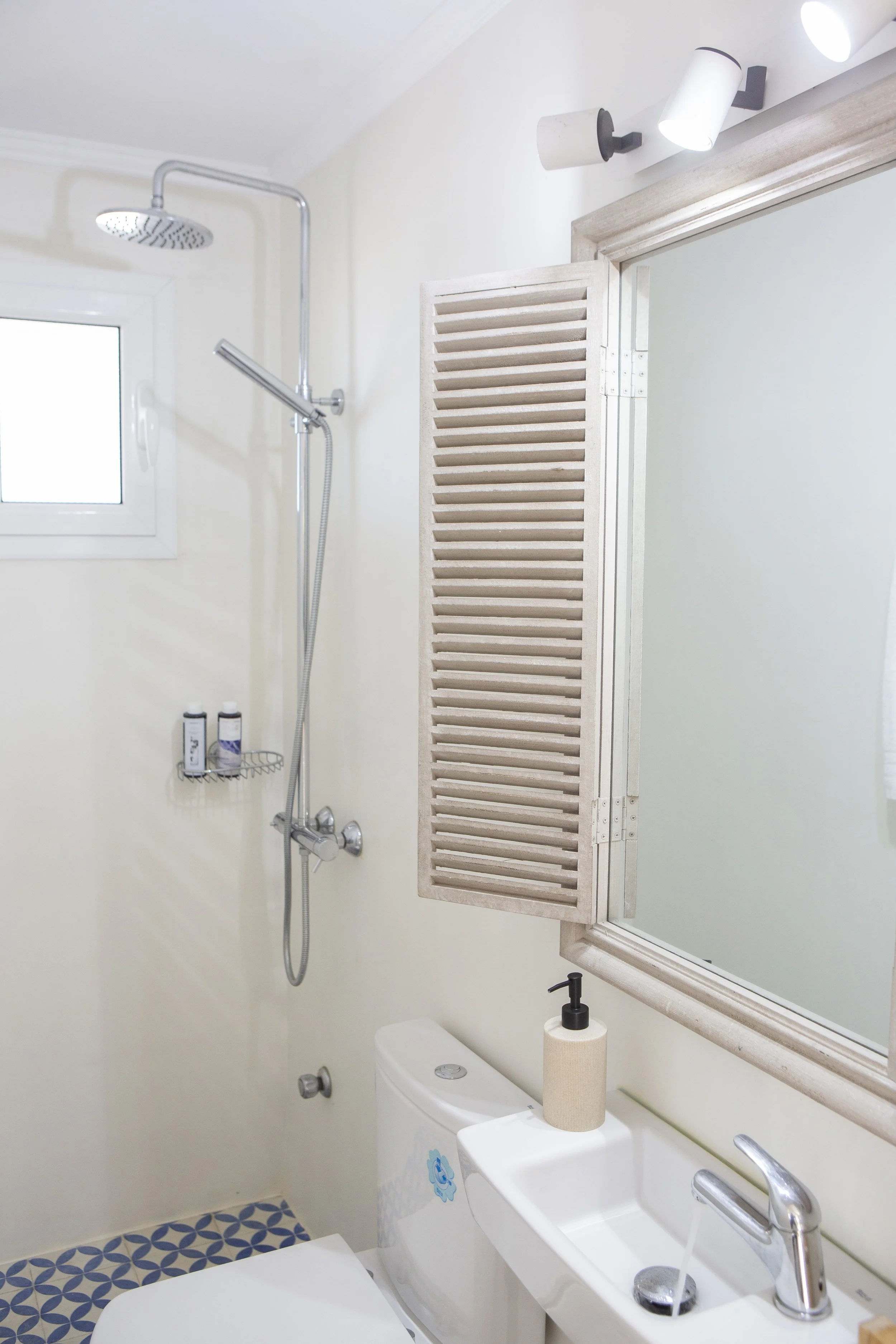 A bathroom with a shower, a mirror, a toilet, a sink, a roll of toilet paper, a soap dispenser, a small window, and a white louvered door, with some toiletries on a small wire basket.