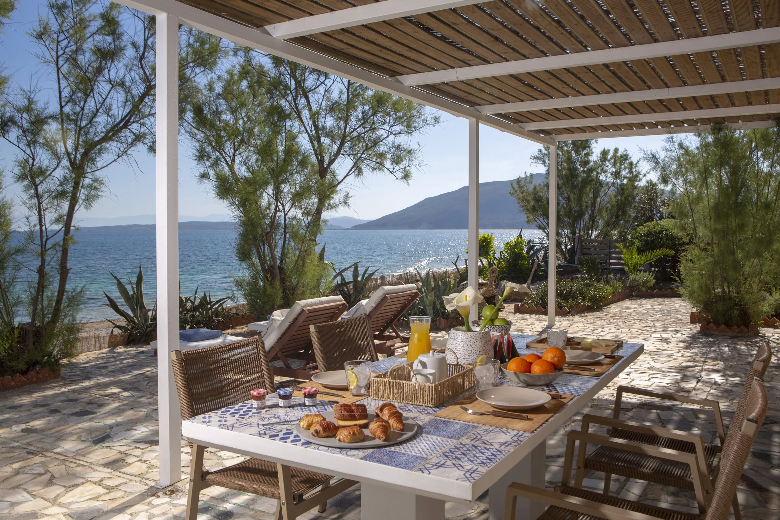 Outdoor patio setup with a table of breakfast foods including pastries, fresh fruit, and drinks, overlooking a lake with trees and mountains in the background.