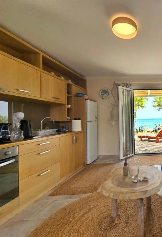 A cozy kitchen with wooden cabinets, a white refrigerator, and a view of a patio with lounge chairs overlooking the ocean through an open glass door.