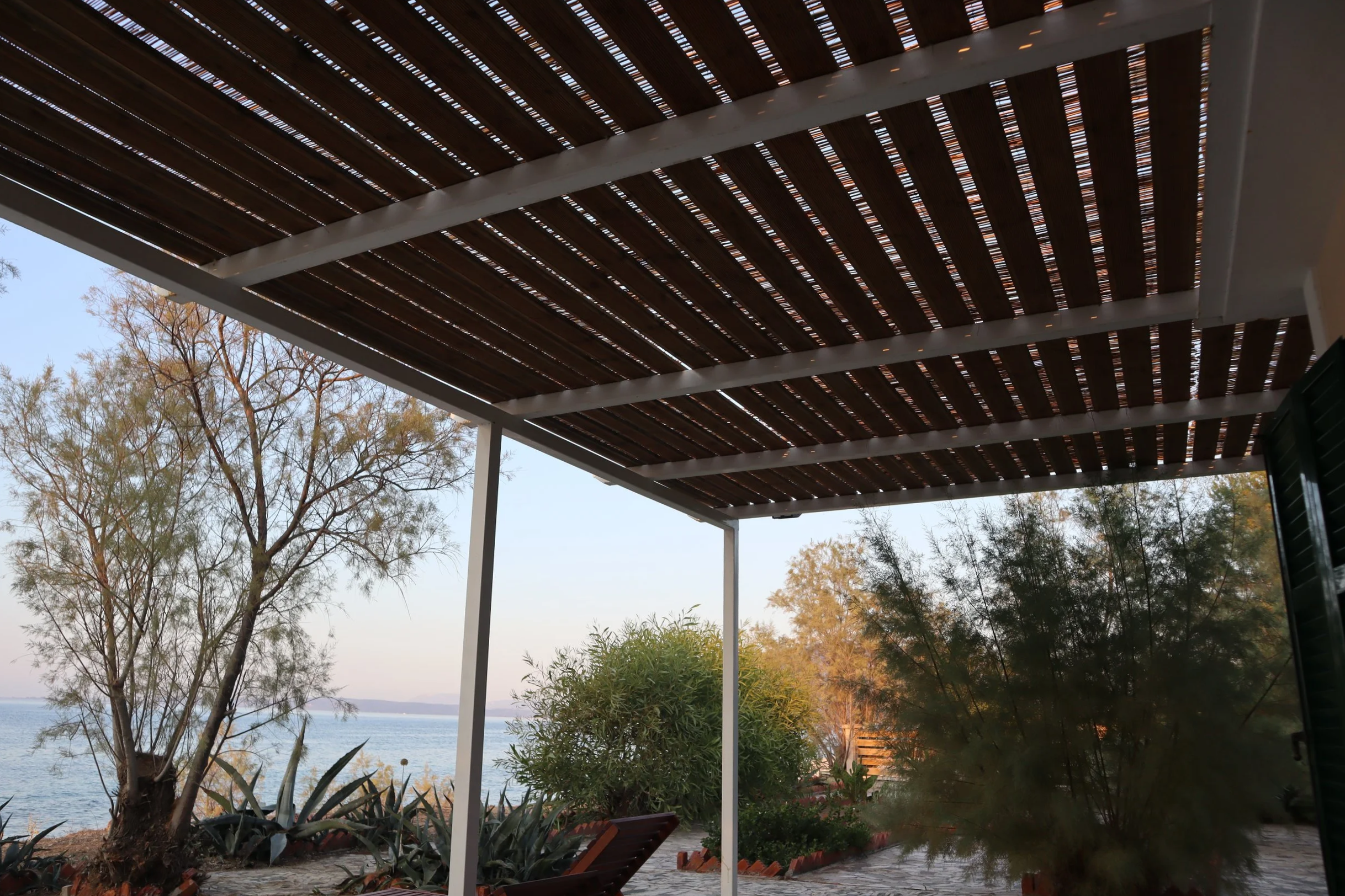 View of a patio with a wooden slat roof, overlooking a body of water, with trees and plants in the background.