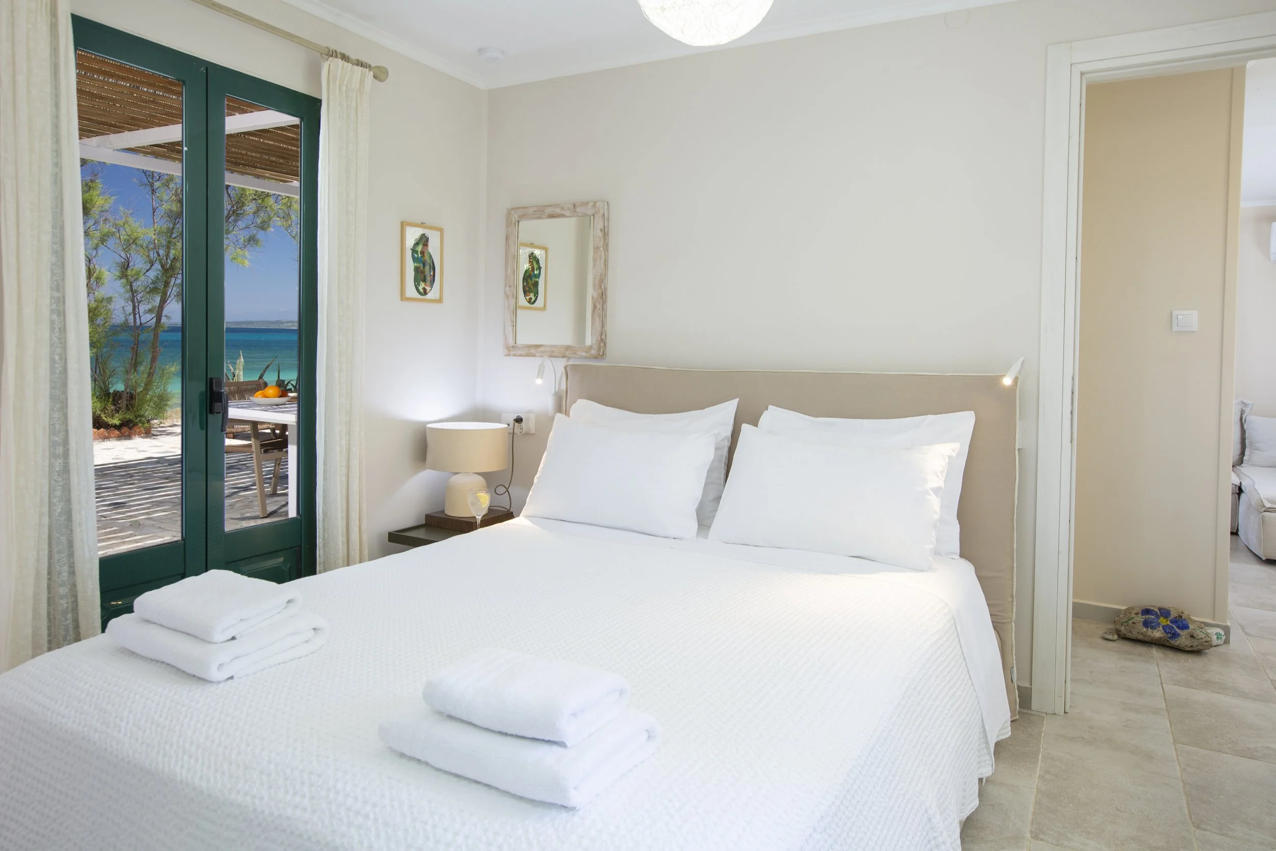 A bedroom with a neatly made bed with white linens, two pillows, and a rolled towel at the foot. There is a nightstand with a lamp and a glass of water next to the bed. The room has a door leading to a balcony with a view of trees, the sea, and a blu