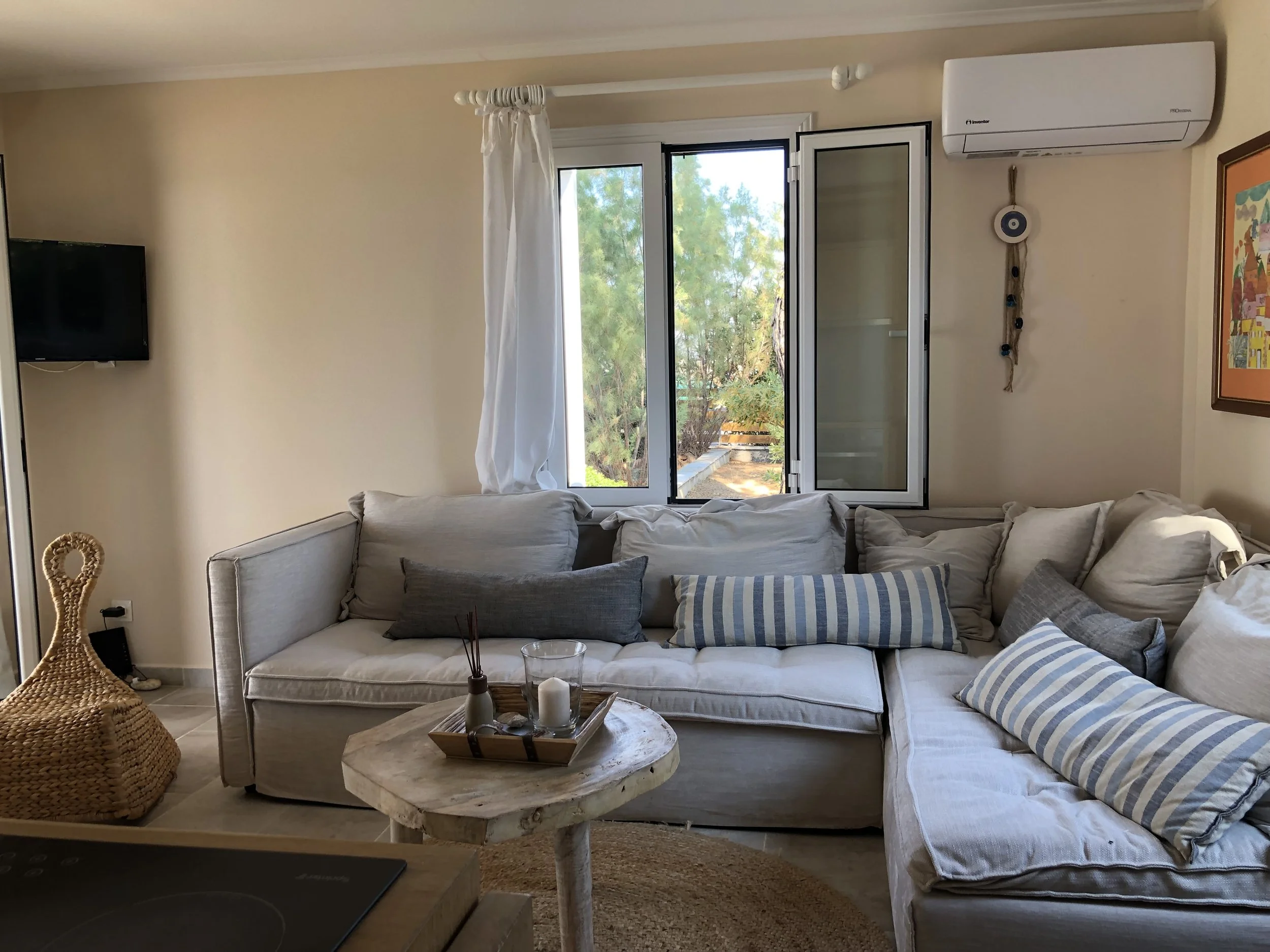 Living room with a beige sectional sofa with various striped and solid decorative pillows, a round wooden coffee table with candles and reed diffusers, a window with white curtains, a wall-mounted TV, an air conditioning unit, a colorful framed pictu