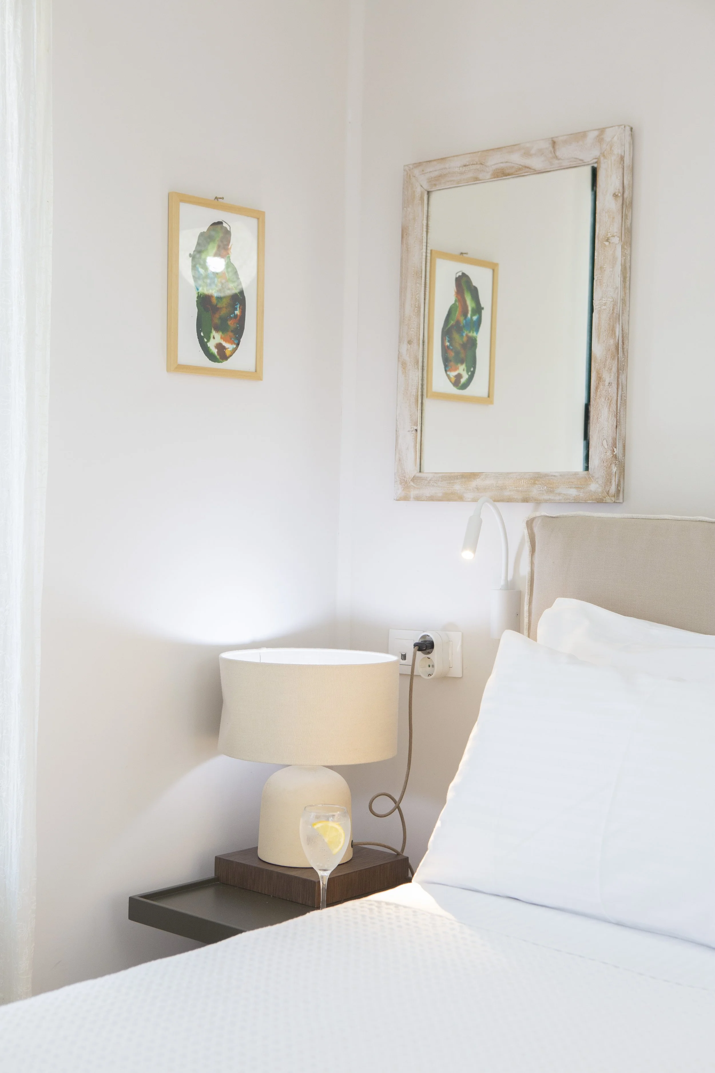 Close-up of a bedroom corner showing a bed with white linens, a beige side table with a beige lamp, and a glass of water with lemon on the table. A mirror with a distressed wooden frame hangs on the white wall, reflecting a framed abstract artwork of
