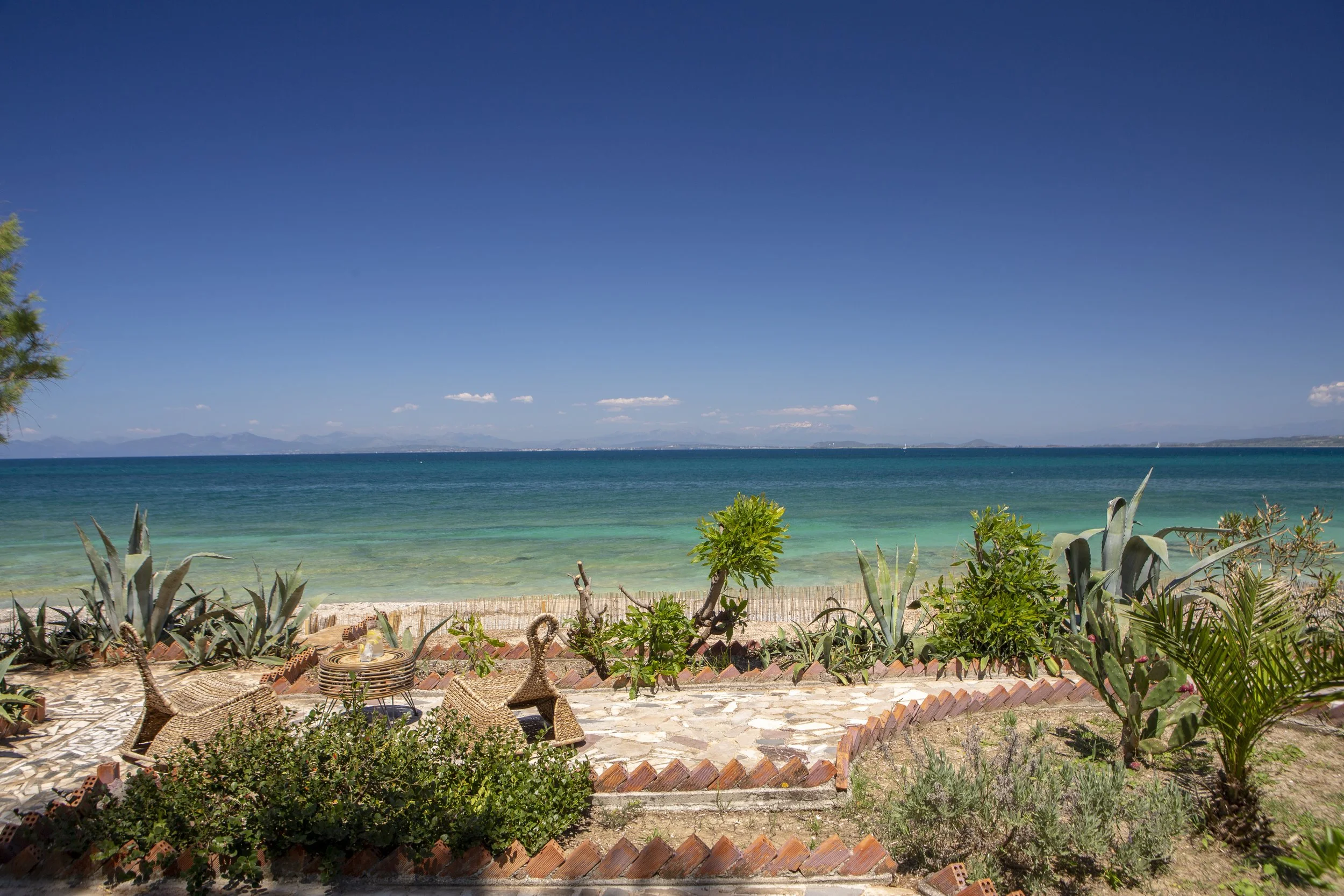 A scenic view of a beach with turquoise water under a clear blue sky, some clouds, and distant mountains on the horizon. In the foreground, there are various green plants, a stone pathway, and outdoor furniture.