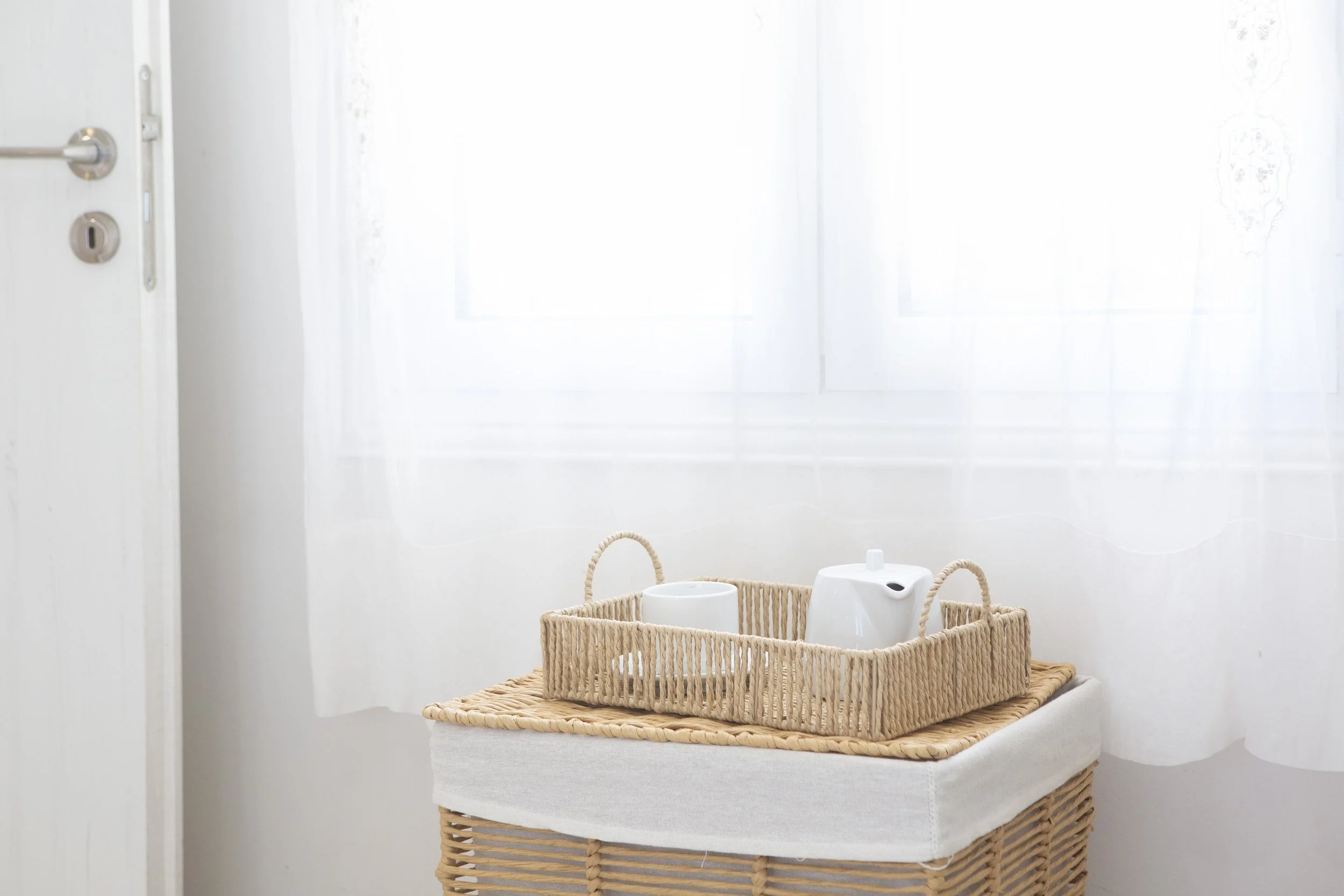 Wicker basket on a table with white ceramic watering cans and a white pitcher, set against a backdrop of a window with sheer curtains.