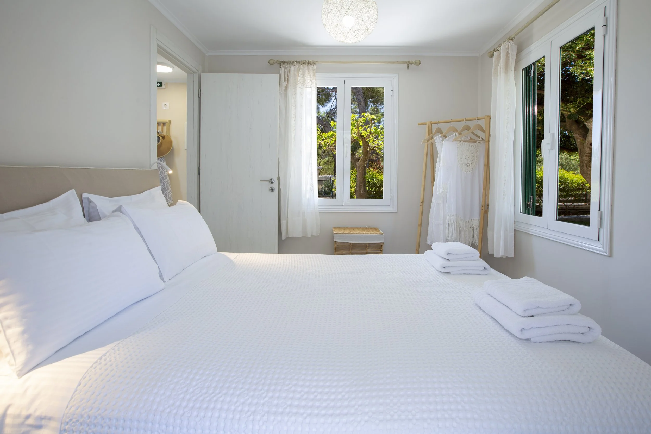 A bright bedroom with white walls, a bed with white linens, and neatly folded towels on the bed. There are two large windows with view of green trees, white curtains, and a clothing rack with white garments. The room is illuminated by natural light a