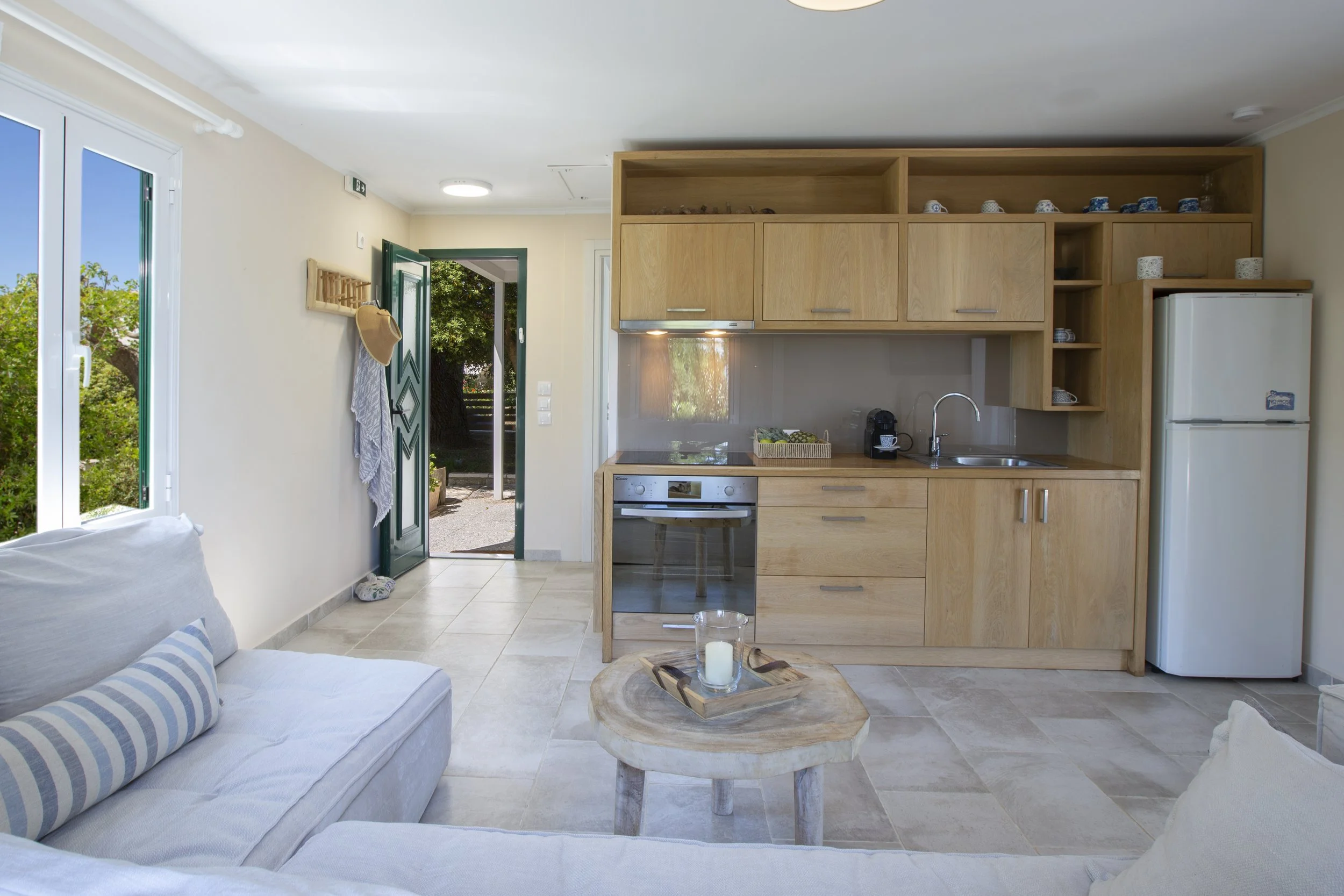 An open-plan living room and kitchen with light-colored tiles, a white sofa with striped cushions, a round wooden coffee table with candles, a kitchen area with wooden cabinets, a stainless steel oven, a coffee maker, a white refrigerator, and a door