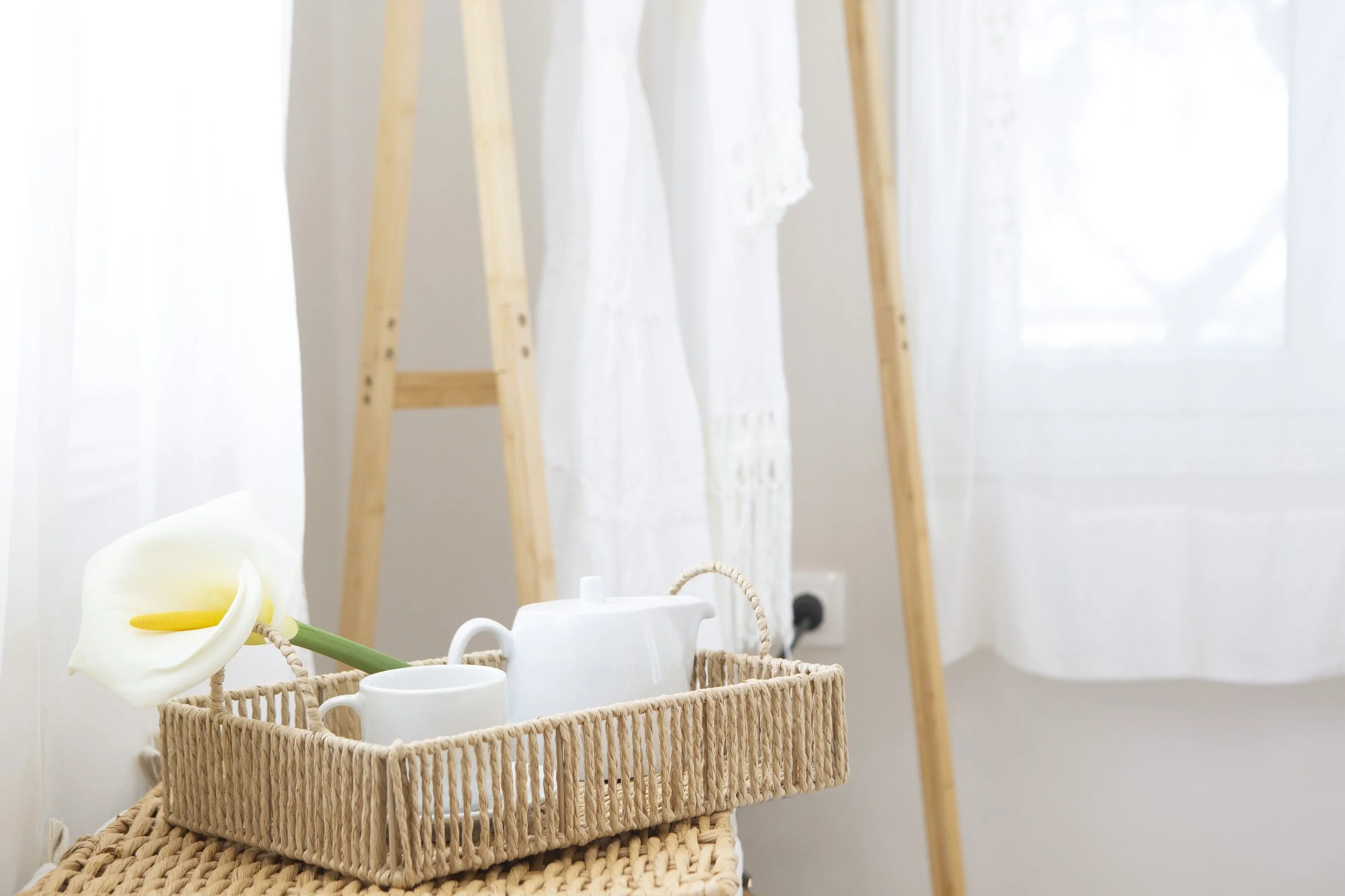 A wicker basket holding a white flower, atop a white tea set with teapot and cups, in front of a wooden ladder and sheer curtains with light filtering through.