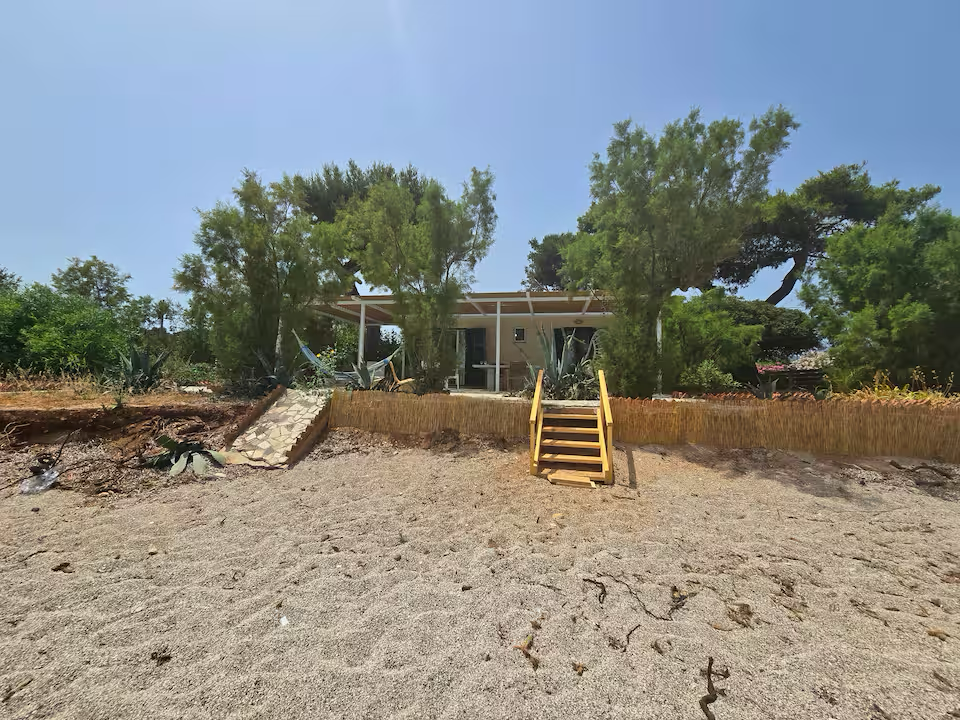 Small house with a porch, surrounded by trees, viewed from a sandy beach with stairs leading up to the house.