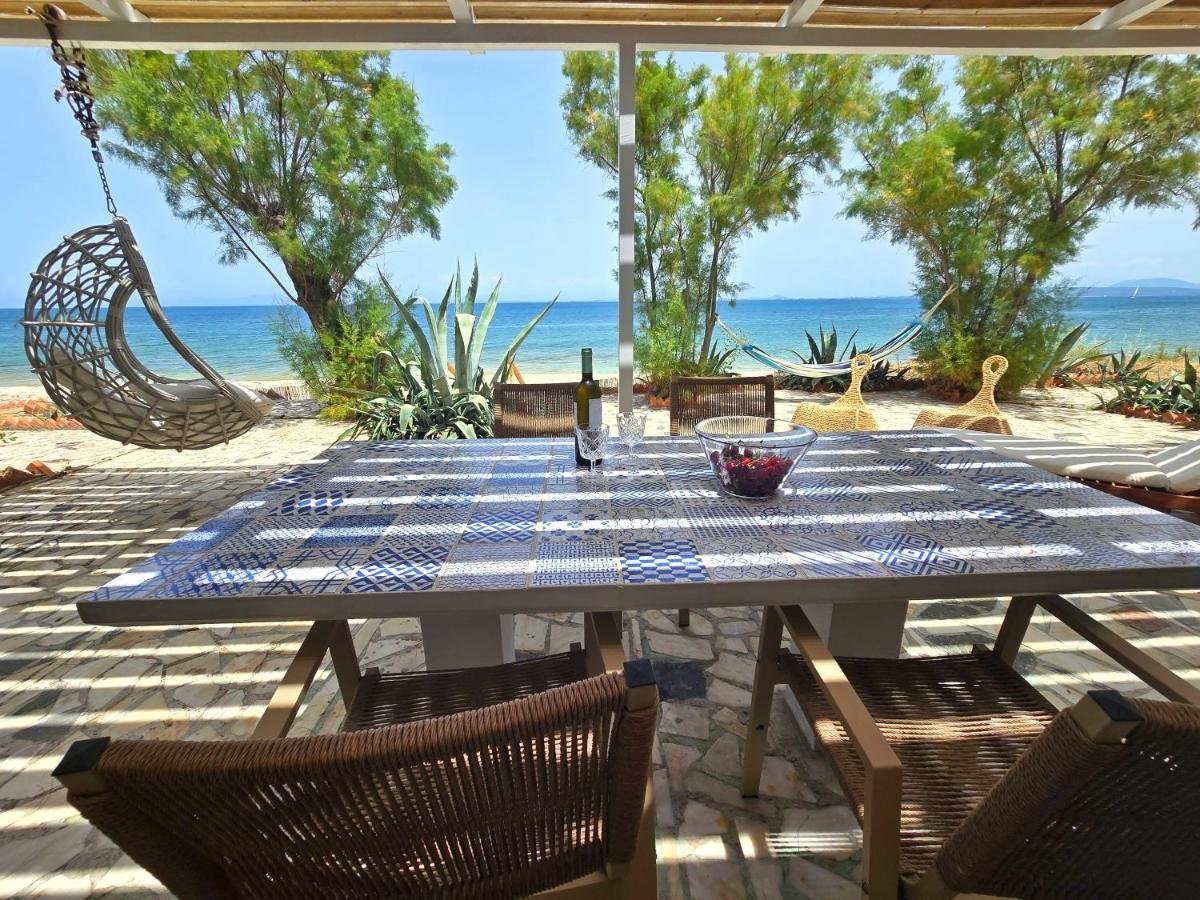 Outdoor dining area on a patio overlooking the beach with a table, chairs, wine, glasses, and a bowl of cherries, shaded by a pergola with a view of the ocean and trees.