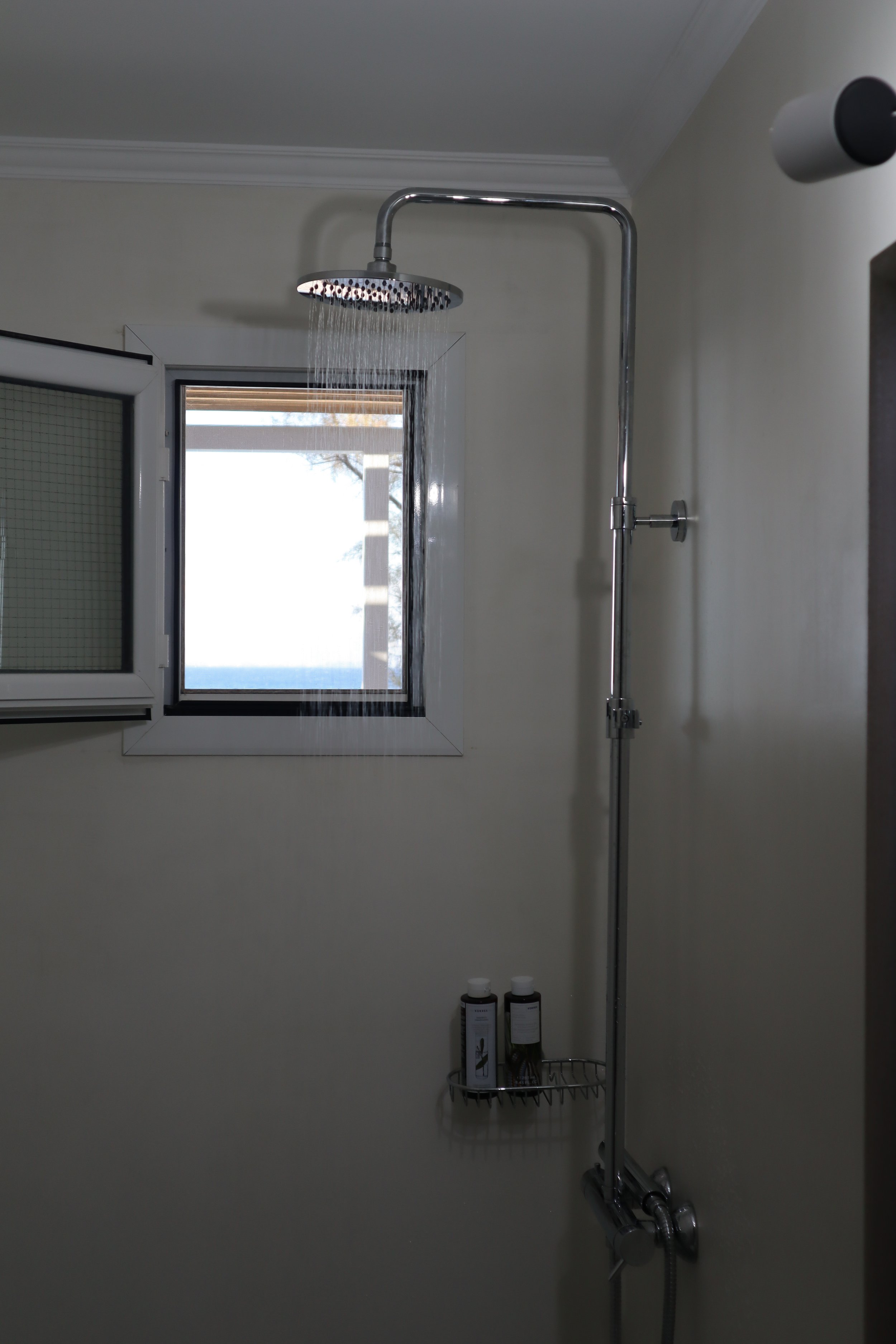 Shower area with a rainfall showerhead, a window, a small shelf with shampoo bottles, and a white wall.