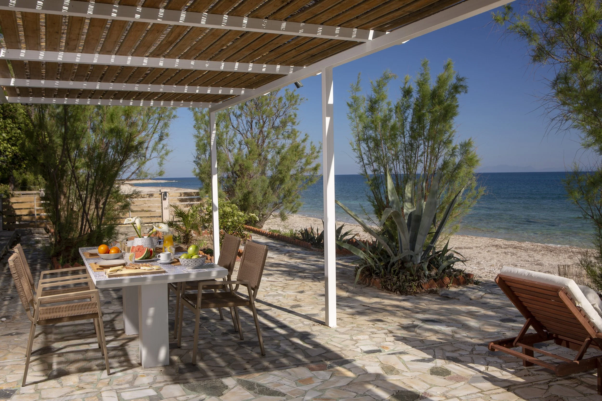 A seaside outdoor patio with a dining table set with fruits, water, and breakfast foods underneath a pergola, with trees, sandy beach, and ocean in the background.