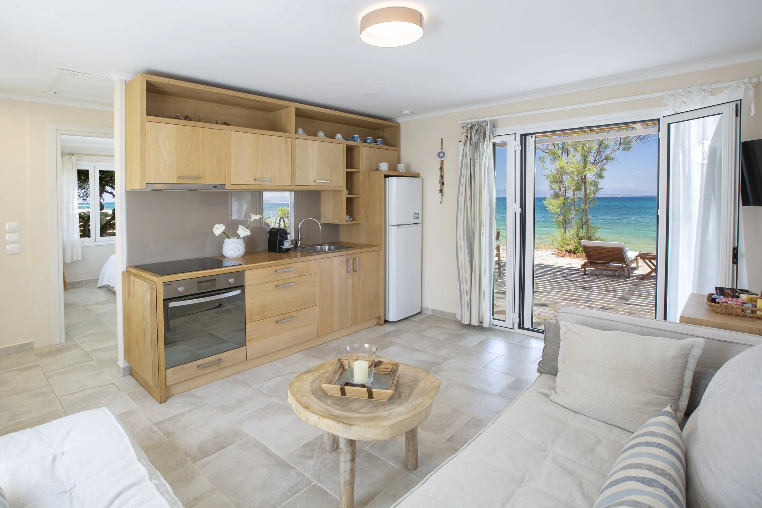 Living room with open sliding glass door showing ocean view, wooden kitchen cabinets, sofa with striped pillow, round wooden coffee table with a candle, and beige tiled floor.