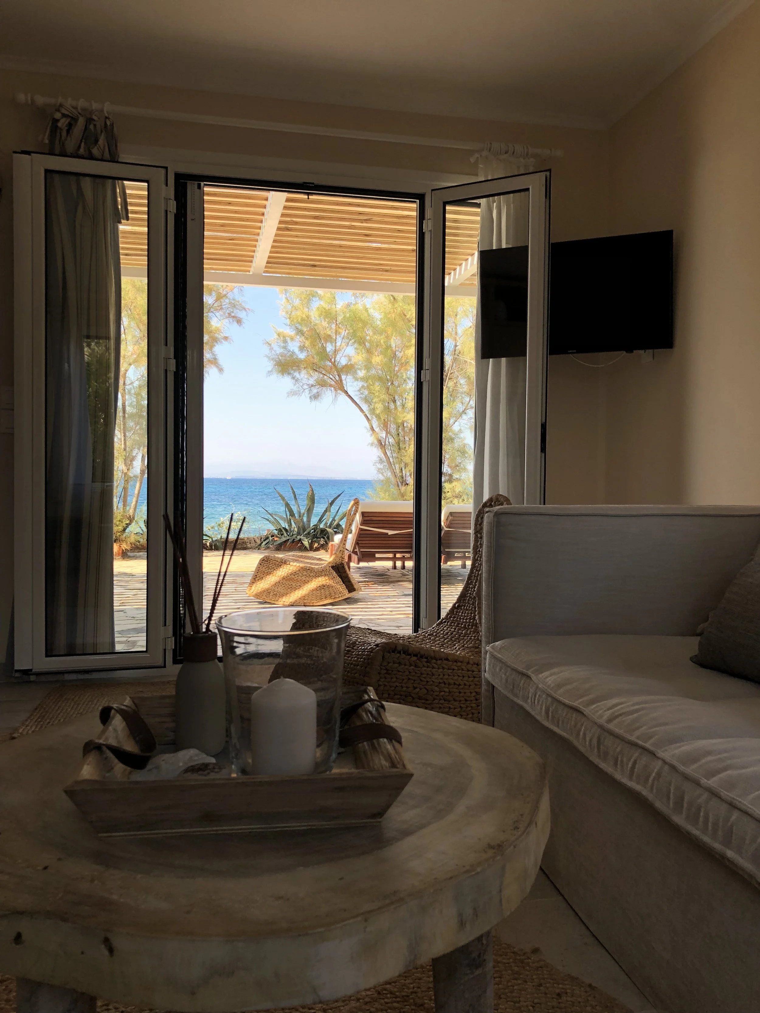 Living room with open glass doors revealing outdoor patio, trees, and ocean view.