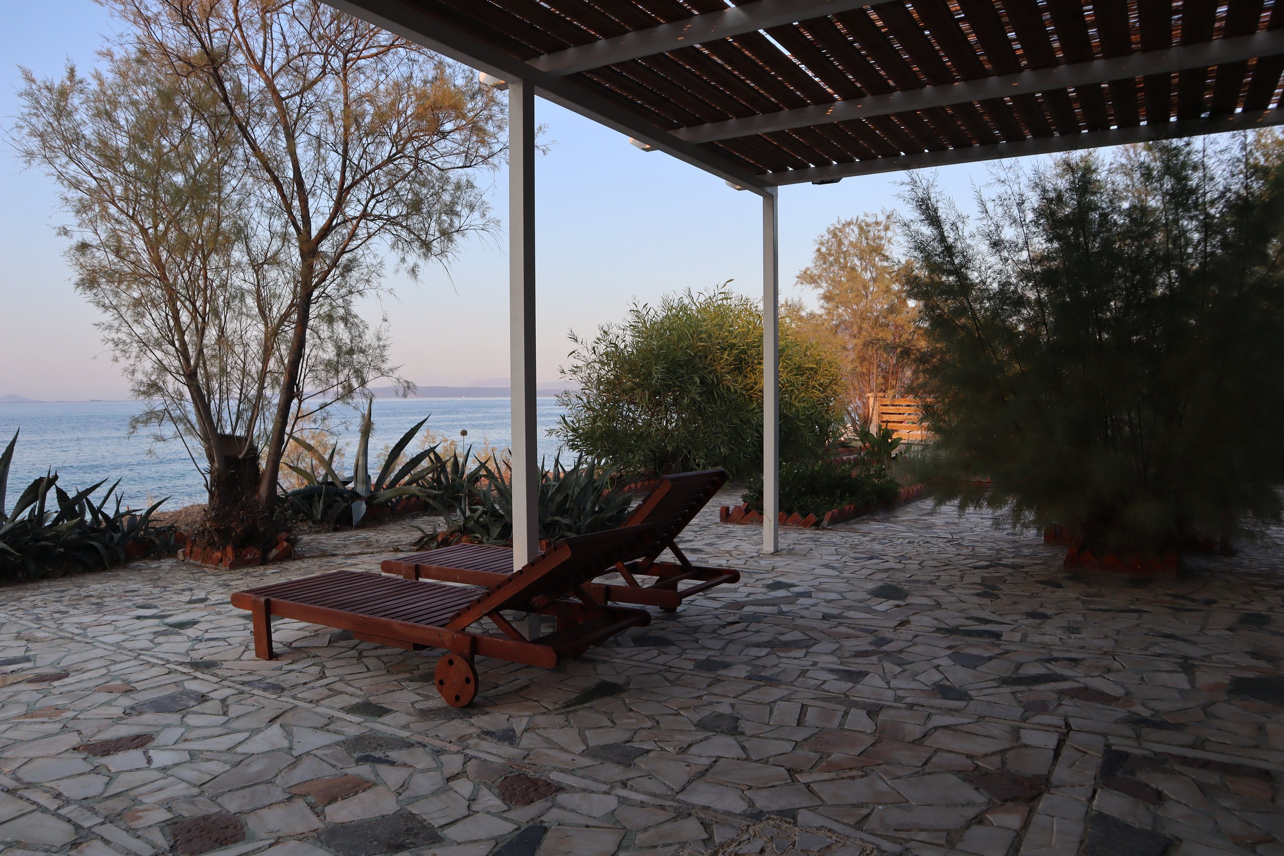 A patio with stone flooring and two wooden lounge chairs, overlooking a body of water with trees and bushes around the edge, under a wooden canopy.
