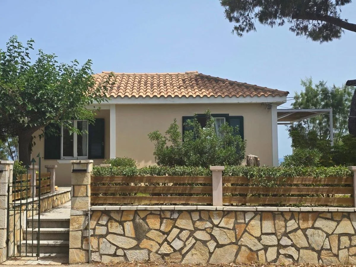 A small house with a beige exterior, black shutters, and a terracotta tiled roof, surrounded by trees and plants, with a stone and wood fence in front.