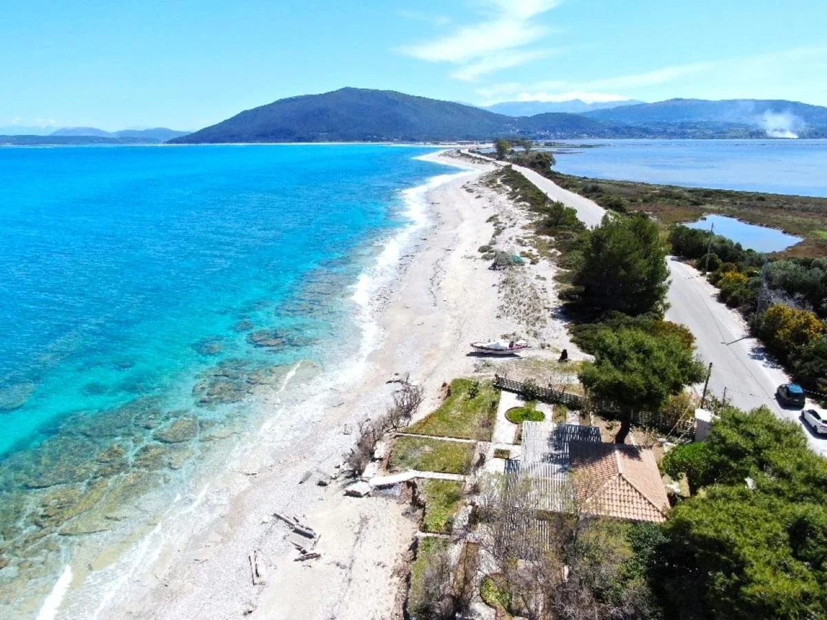 Aerial view of a coastal area with turquoise ocean water, a sandy beach, a road parallel to the coast, and a small house with trees in the foreground. Hills and mountains are visible in the background.