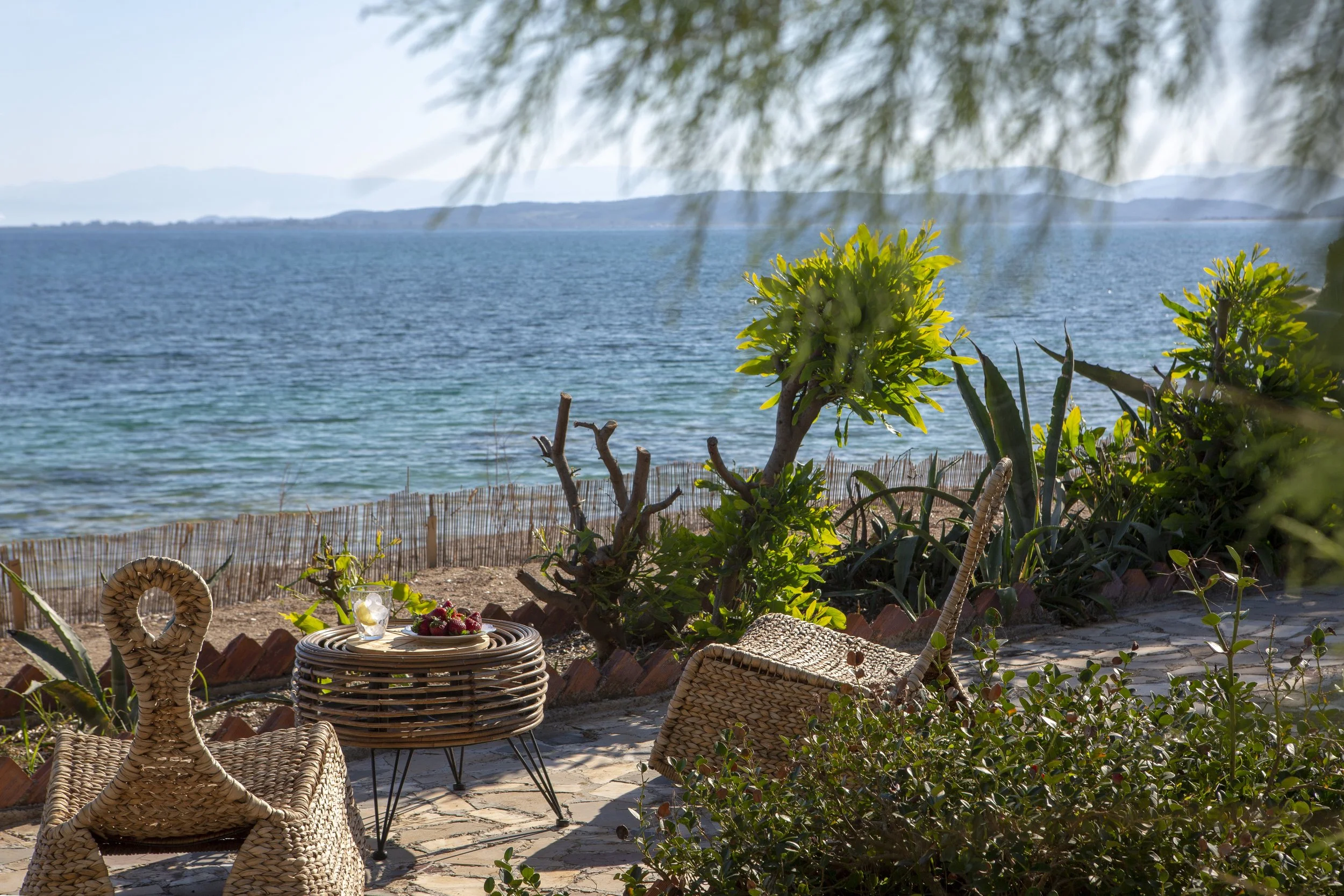 A seaside outdoor patio with wicker chairs, a basket table with strawberries, and a scenic view of the ocean, mountains, and trees in the background.