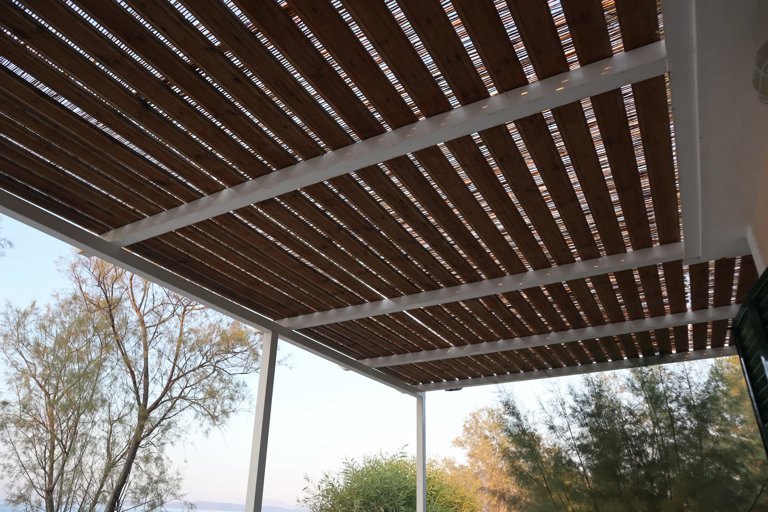 View of a wooden pergola ceiling with trees and sky in the background.