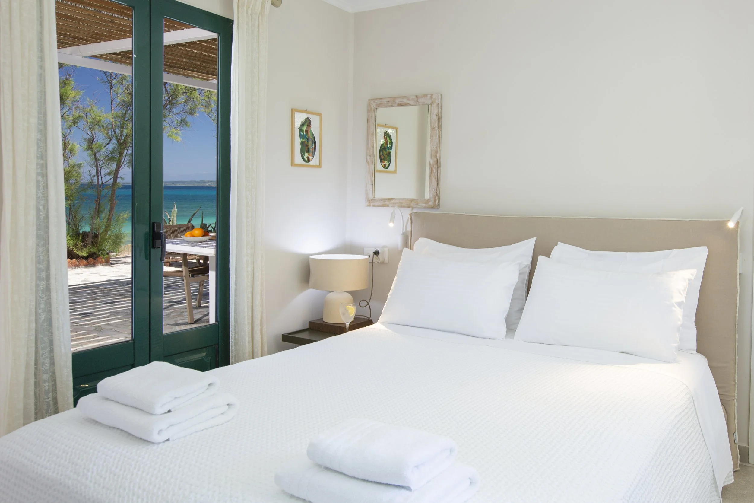 A bright hotel room with a bed covered in white linens and folded towels, a small bedside table with a lamp, and a wall mirror. A sliding glass door opens to a patio with outdoor seating and views of the ocean and trees.