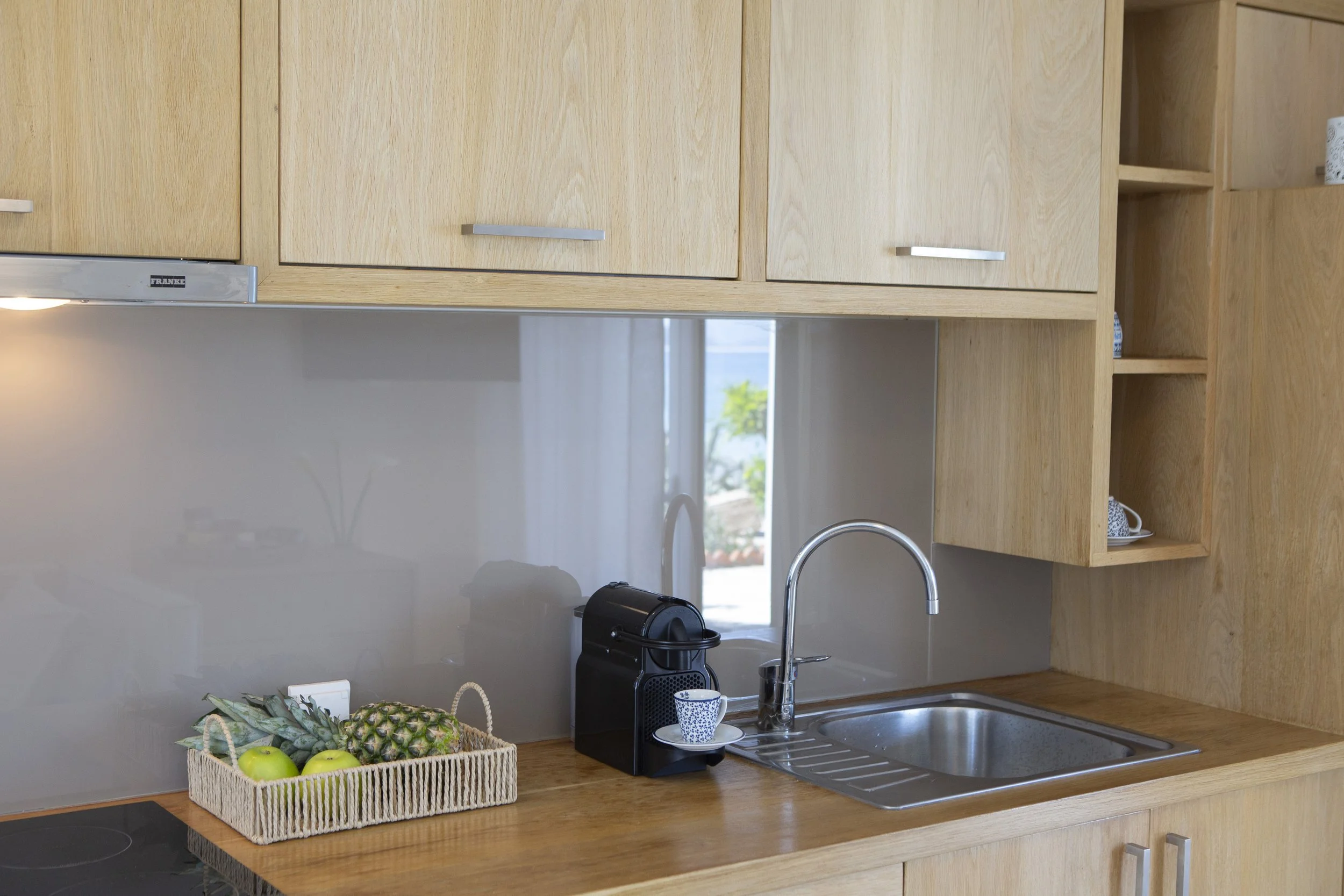 Kitchen countertop with a basket of fruits including pineapples, apples, and bananas. There is a coffee machine and a mug near the sink, with wooden cabinets above.