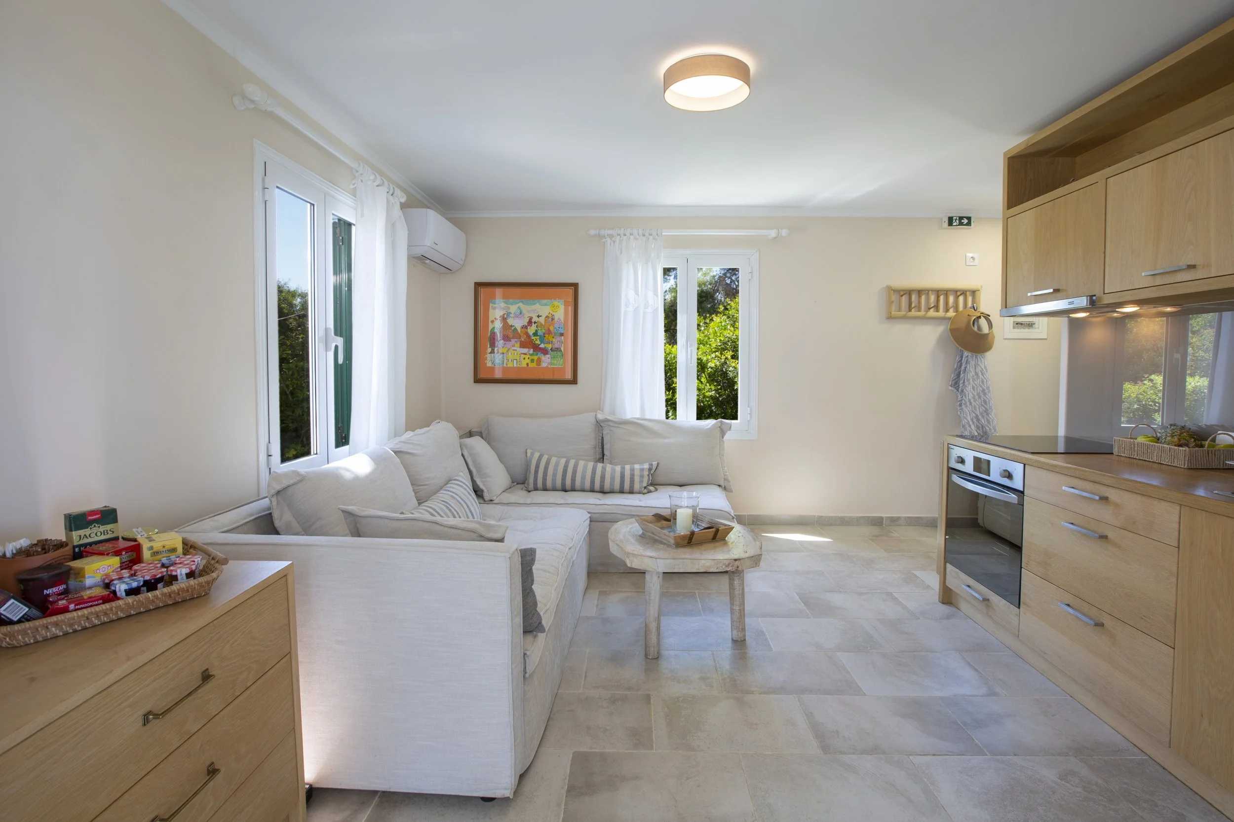 A bright and cozy living room with white walls, a beige sofa with striped pillows, and large windows with white curtains. There is a colorful framed picture on the wall, a small wooden table with a candle, and a light-colored wooden kitchen unit with