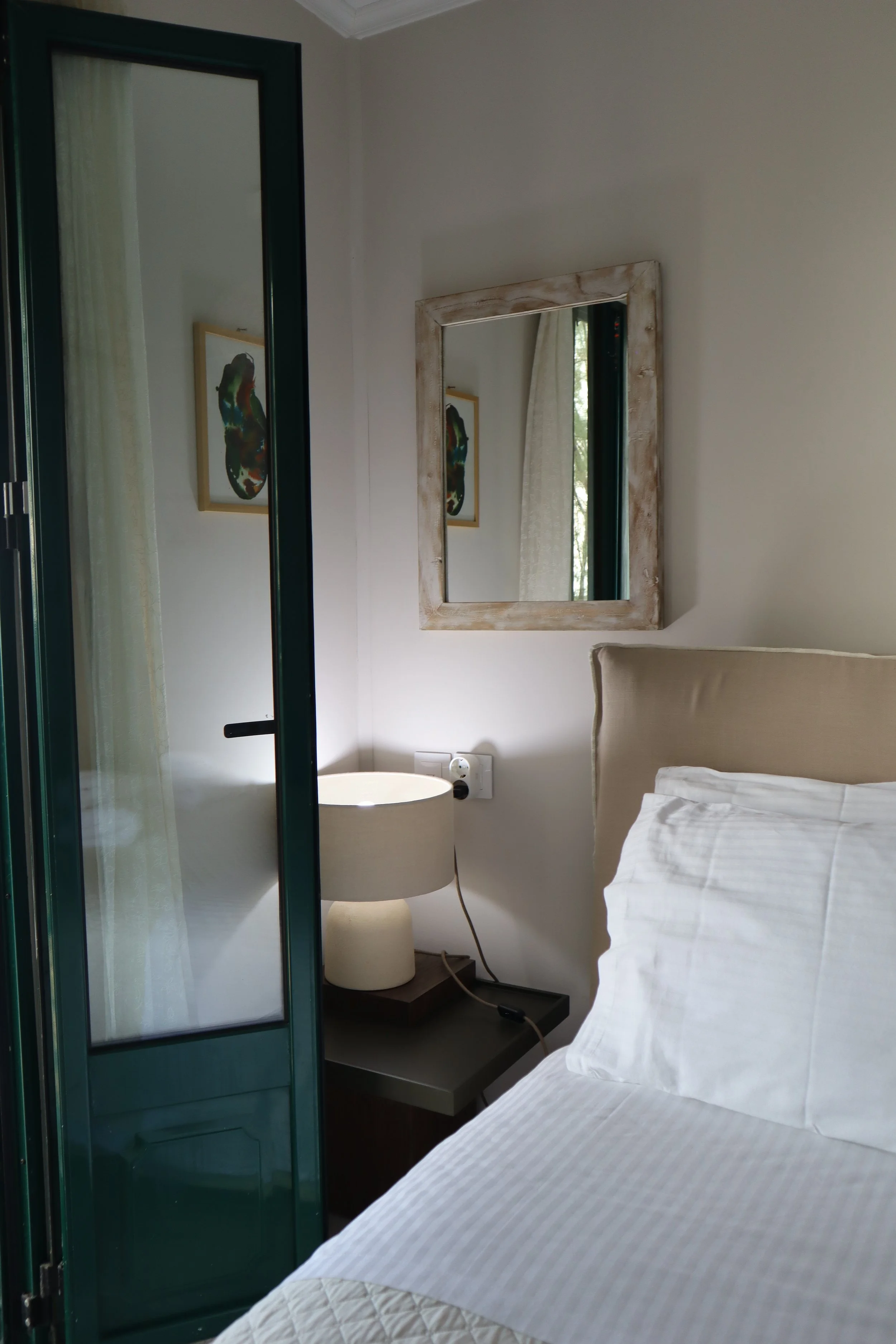 A cozy bedroom corner with a bed, a bedside table with a lamp, a mirror on the wall, and a window with light curtains.